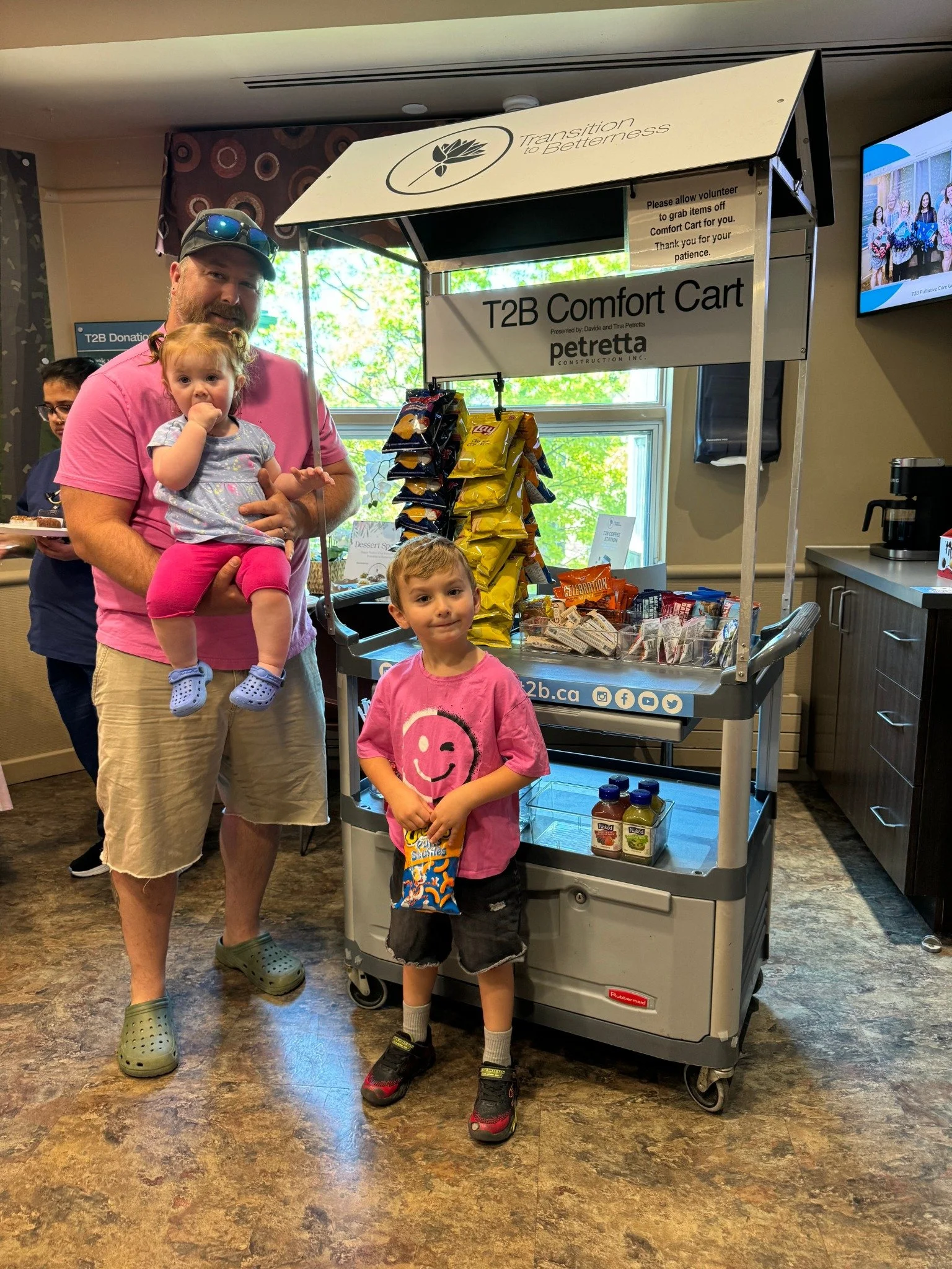 A man holding a young girl with red hair, next to a young boy with brown hair, standing in front of a snack cart labeled 'T2B Comfort Cart'. The man is wearing a pink T-shirt and beige shorts, the girl is wearing a light blue top and pink pants, and the boy is wearing a pink T-shirt with a smiley face and black shorts, holding a snack. The cart has chips, snacks, and drinks, with a window behind it showing green trees outside.