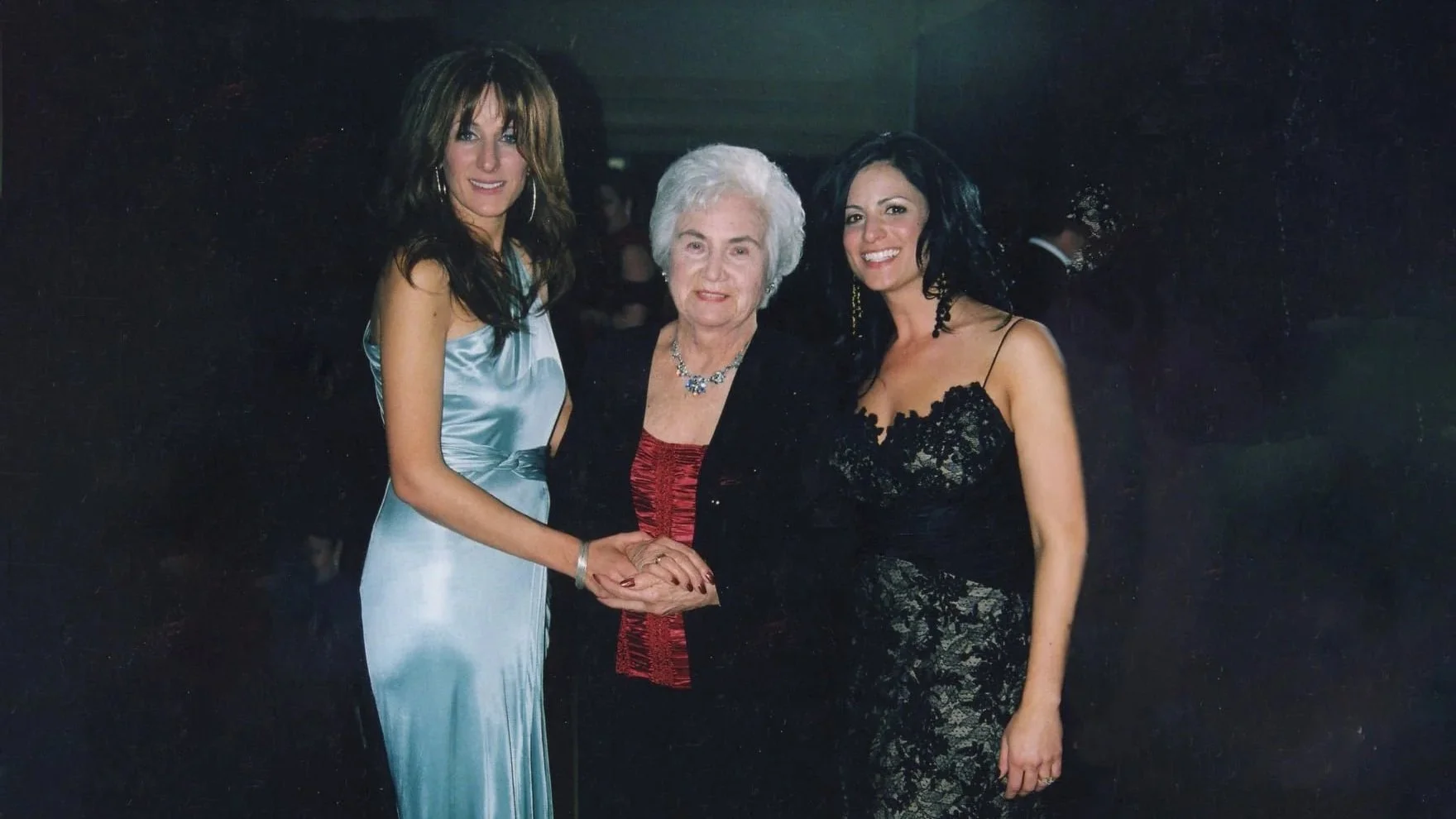 Three women standing together at an indoor event, holding hands and smiling at the camera. The woman in the middle is older with white hair. The women on the sides are younger, wearing elegant dresses.