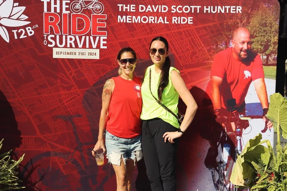 Two women standing in front of a red event poster for 'The Ride to Survive,' promoting the David Scott Hunter Memorial Ride scheduled for September 21, 2024. One woman wears a red sleeveless top, sunglasses, and ripped denim shorts, holding a drink. 