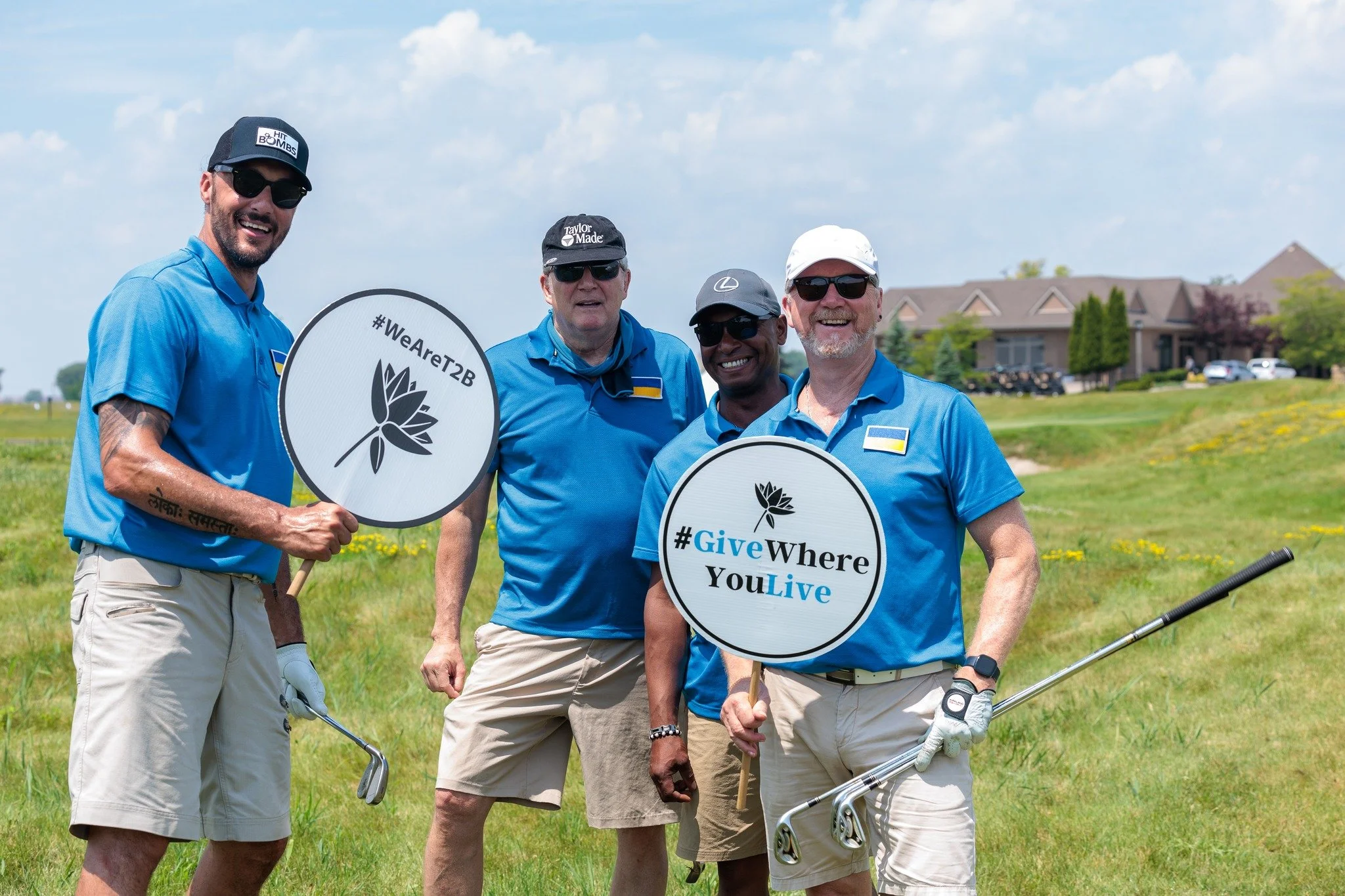 Four men standing on a golf course holding signs with hashtags and a flower symbol, dressed in blue shirts and khaki shorts, smiling at the camera.