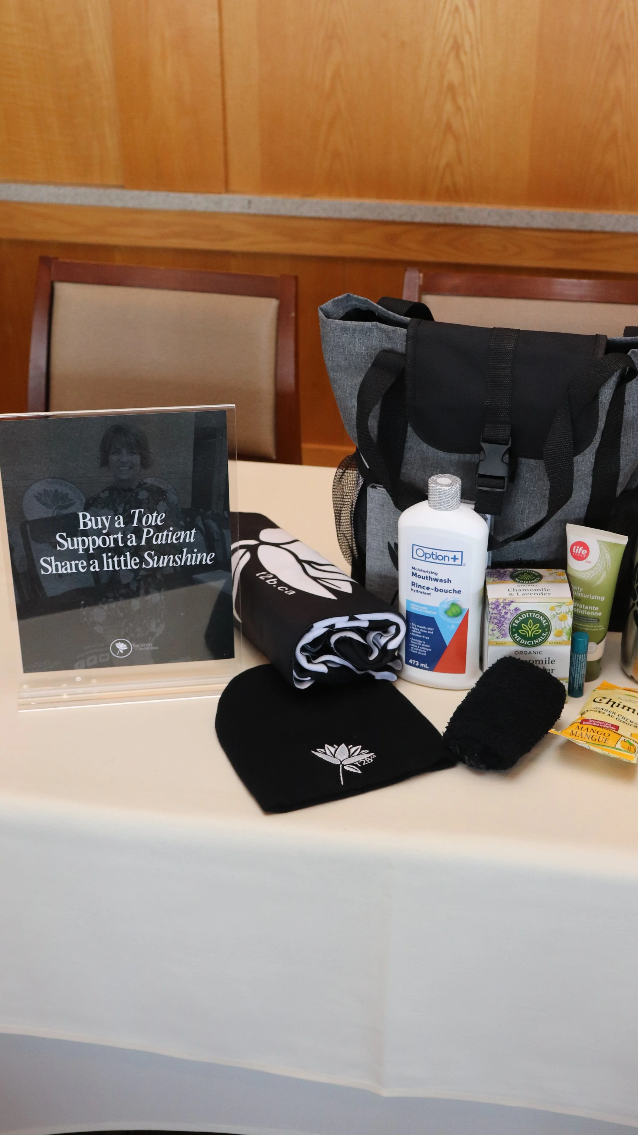 Table with health and personal care items including a bottle of mouthwash, an eye balm, herbal tea, a lip balm, a black beanie with a logo, a small black towel, and a tote bag, with a sign promoting support for patients.
