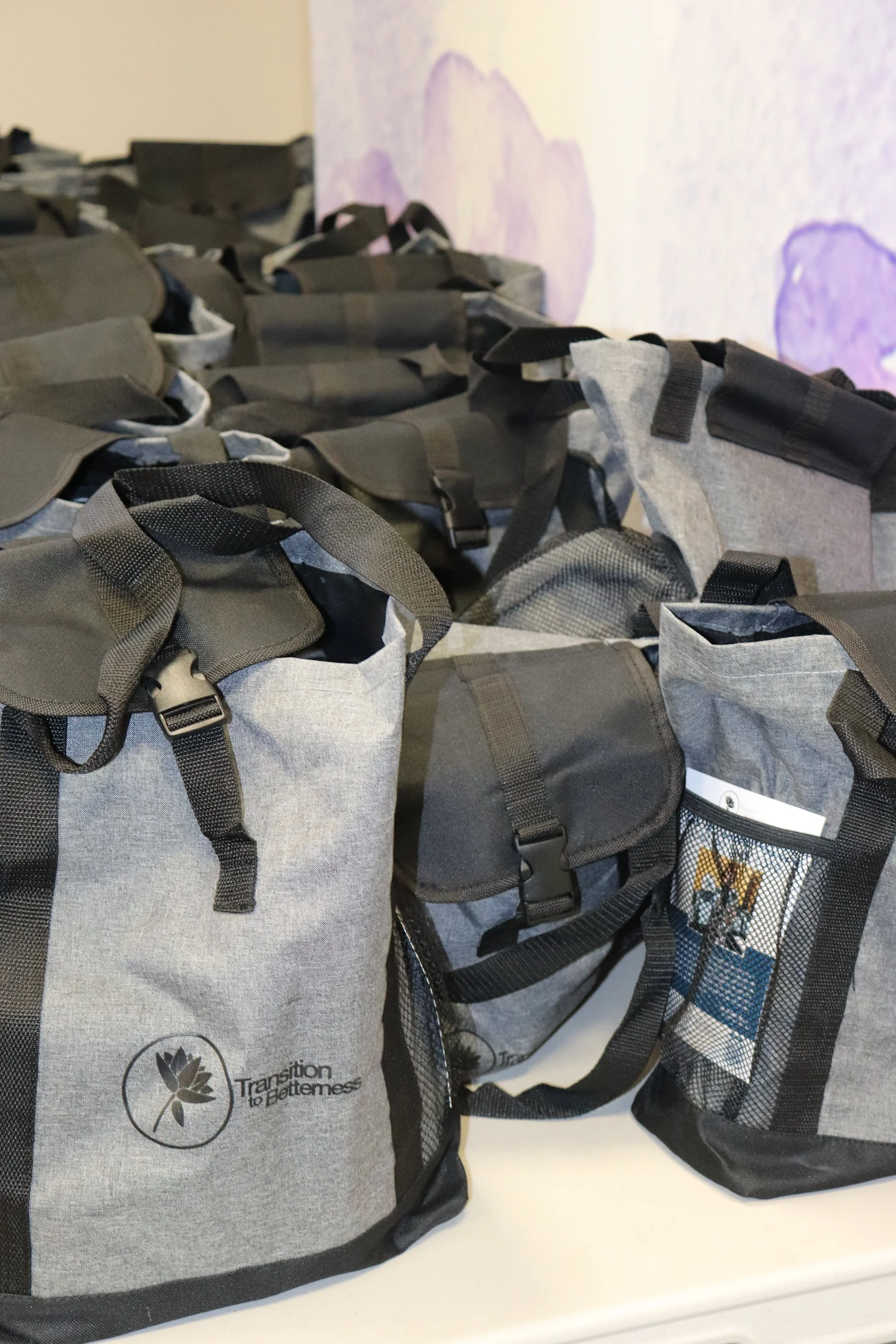 Multiple gray and black backpacks lined up on a white surface, with a purple and white wall in the background.