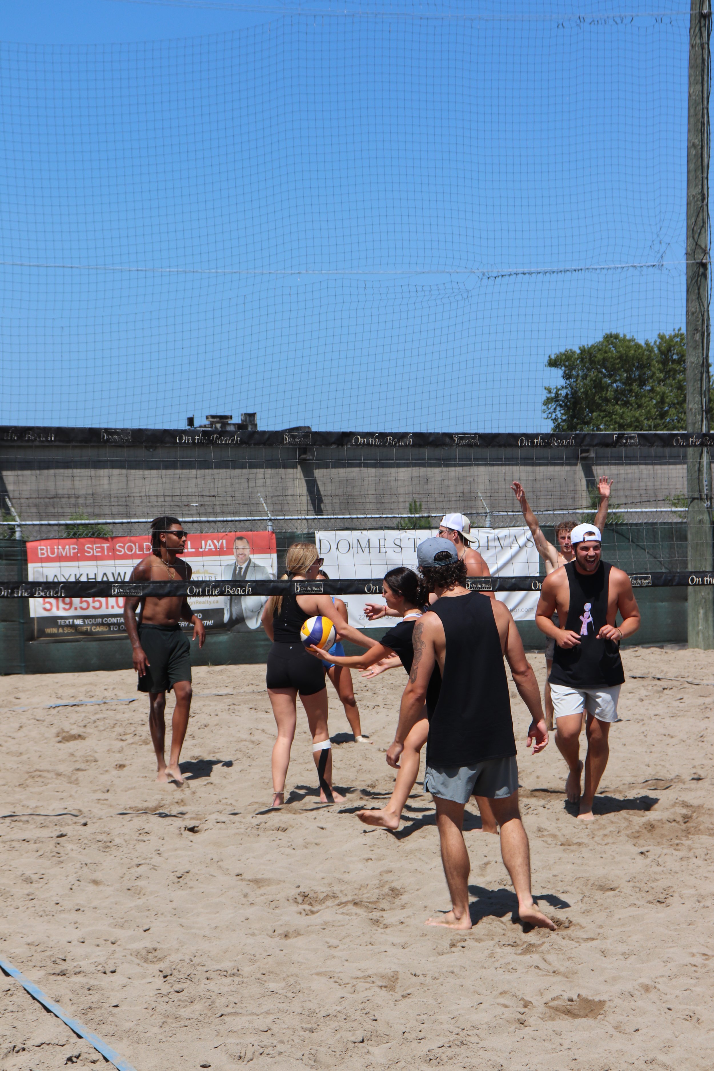 Group of young adults playing beach volleyball on sandy court, some wearing sleeveless shirts and shorts, with blue sky and trees in the background.
