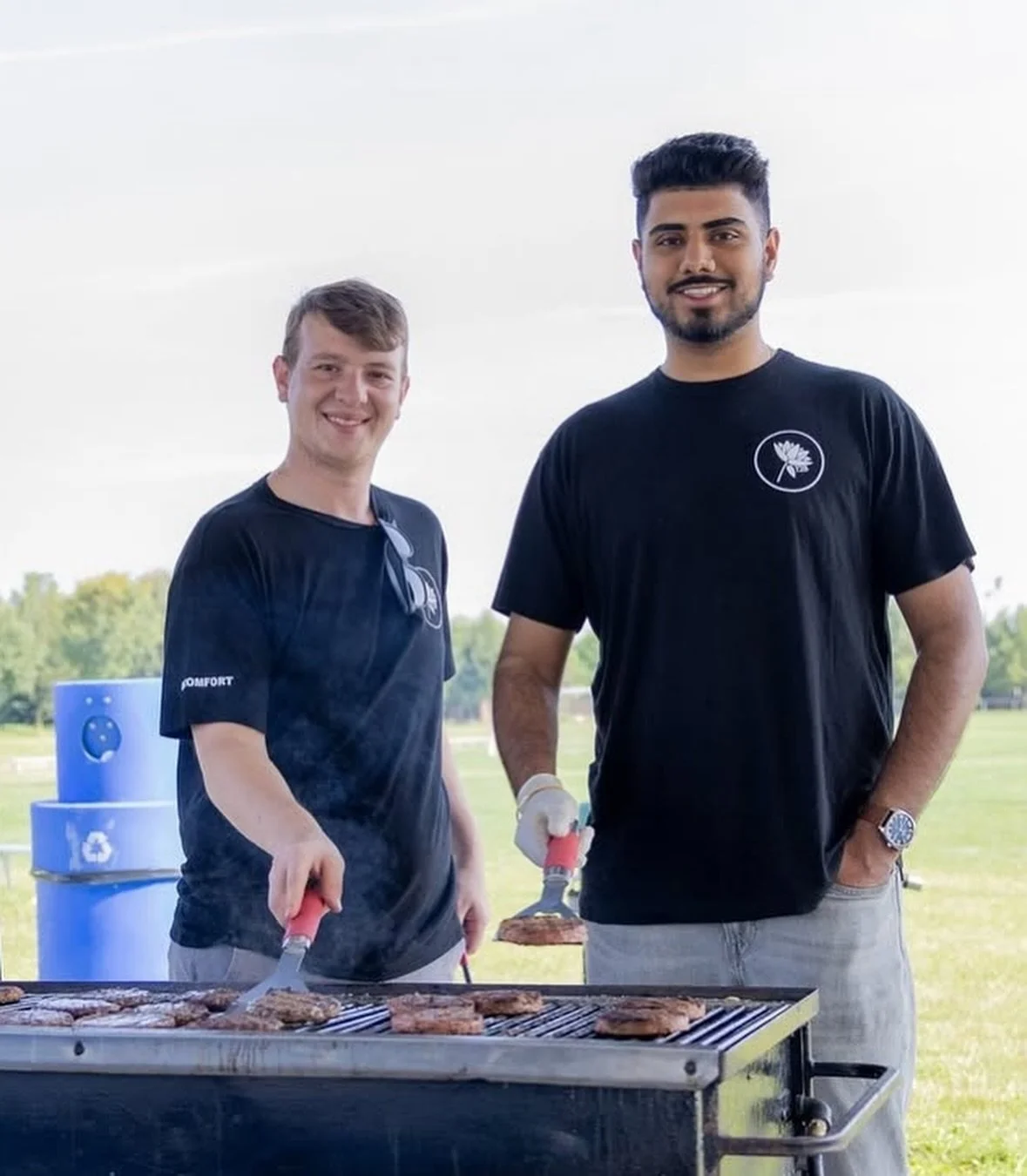 Two men standing outdoors at a barbecue grill, cooking burgers, with a park in the background.