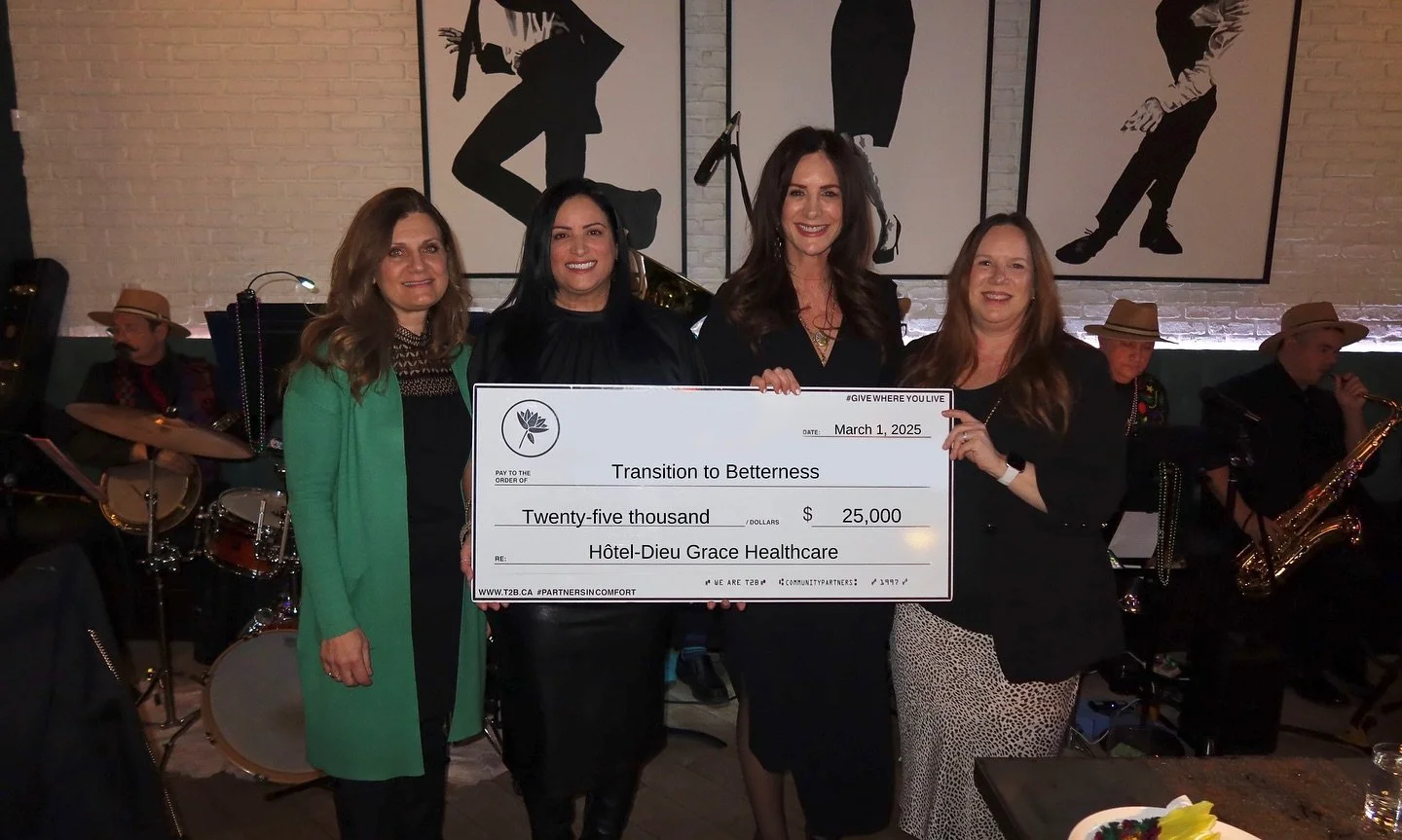 Four women holding a large check in front of a live band playing in a restaurant or bar. The women are smiling and dressed in business or semi-formal attire.