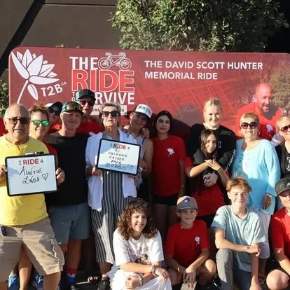 Group of people gathered outdoors in front of a banner for the David Scott Hunter Memorial Ride, some holding signs supporting the event.