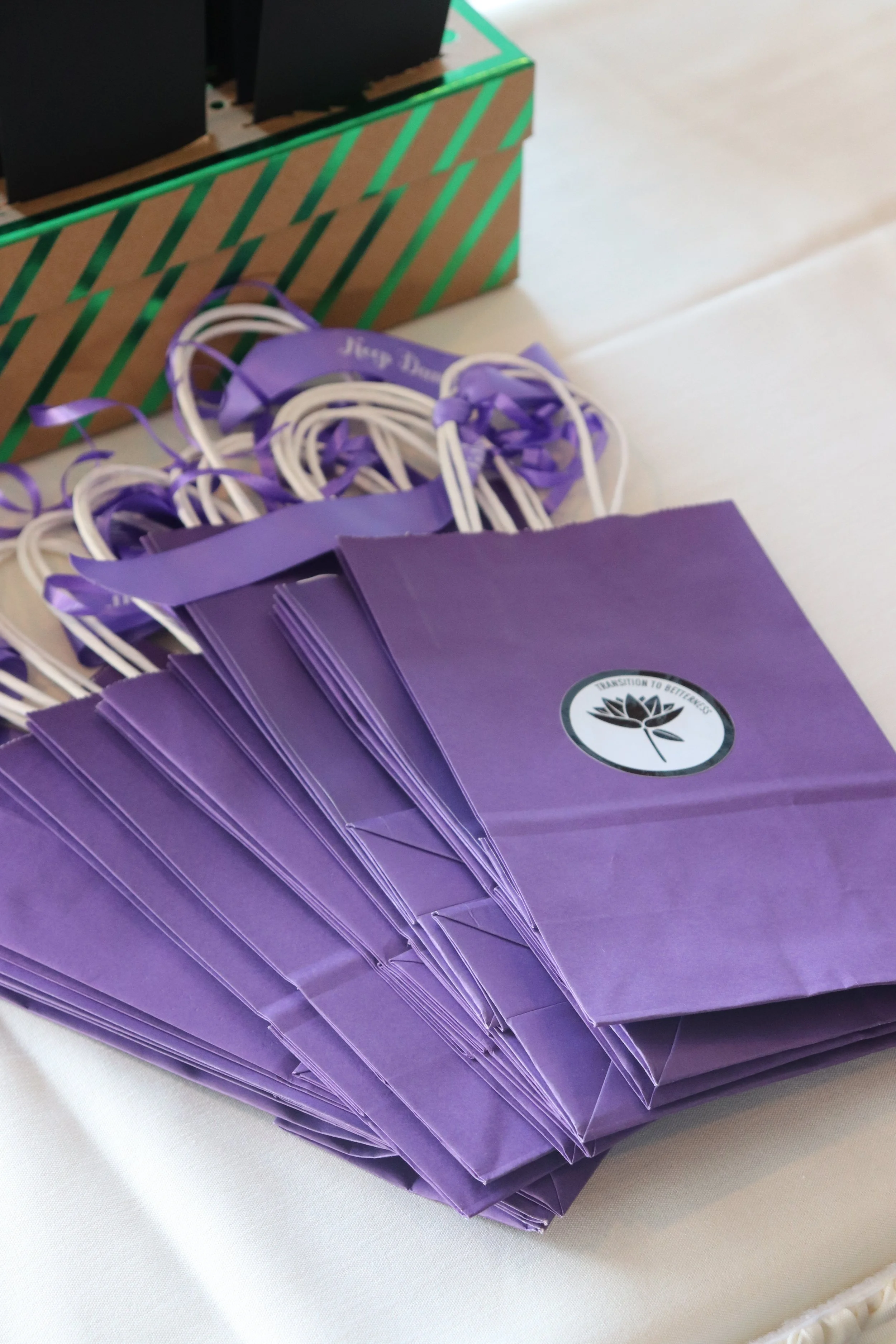 A stack of purple paper gift bags with a circular logo reading 'Transition to Bettering' and a black lily inside the circle, placed on a white surface.