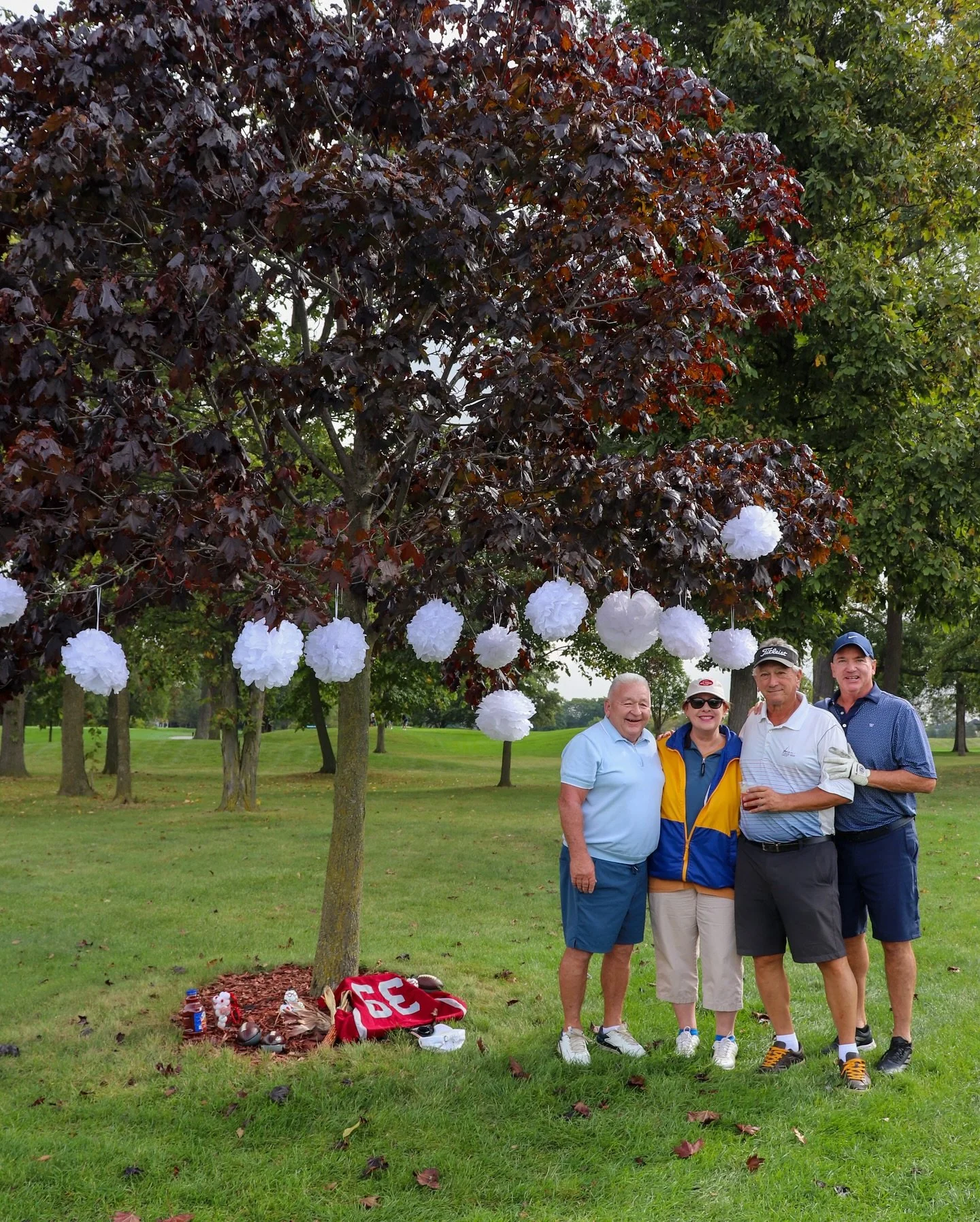 Four people standing under a tree decorated with white pom-pom decorations at a park, with a pile of sports equipment and a bag on the ground nearby.
