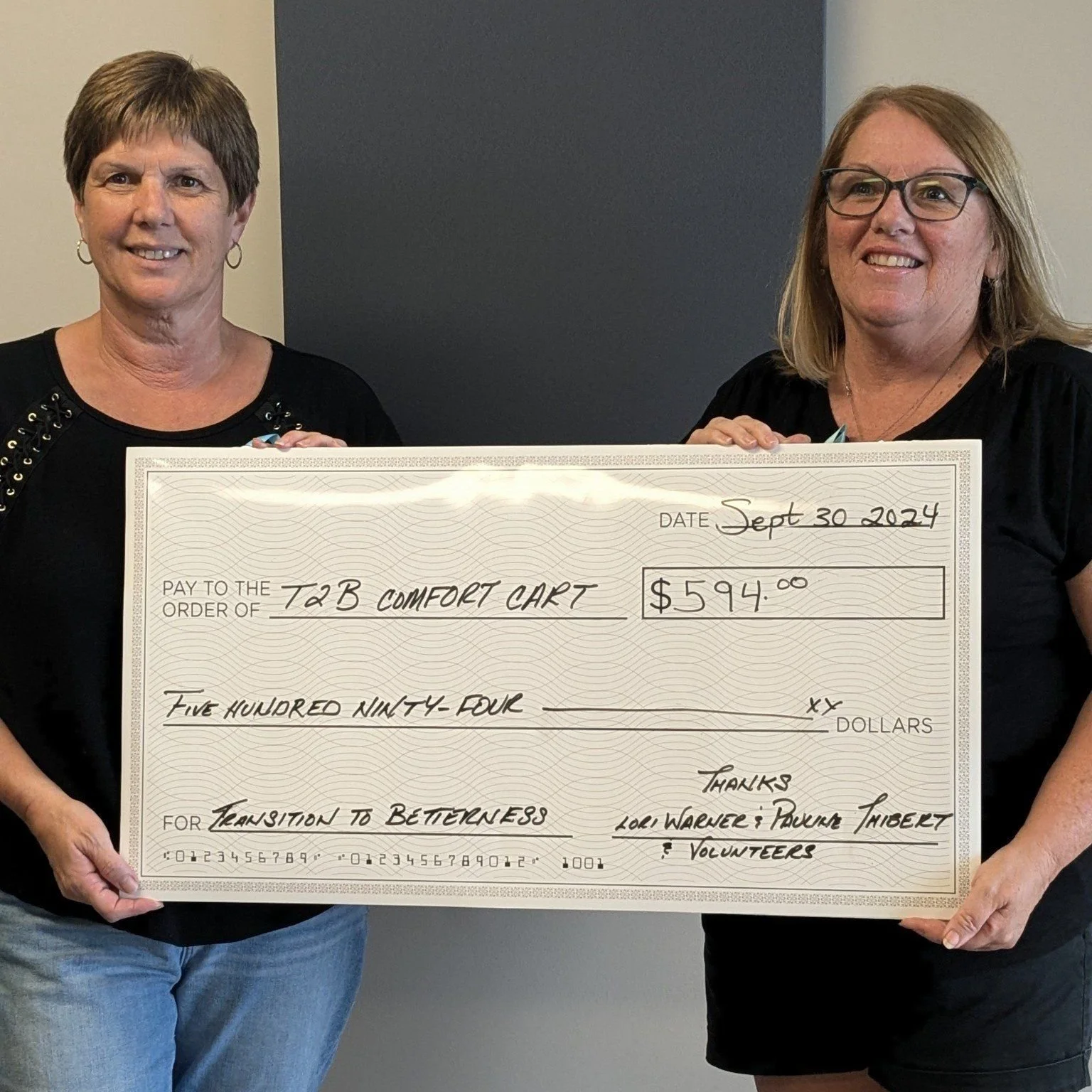 Two women holding a large check for $594.00 made out to T2B Comfort Cart, dated September 30, 2024, with the memo for 'Transition to Brightness' from Lore Warner, Power Thibert, and volunteers.