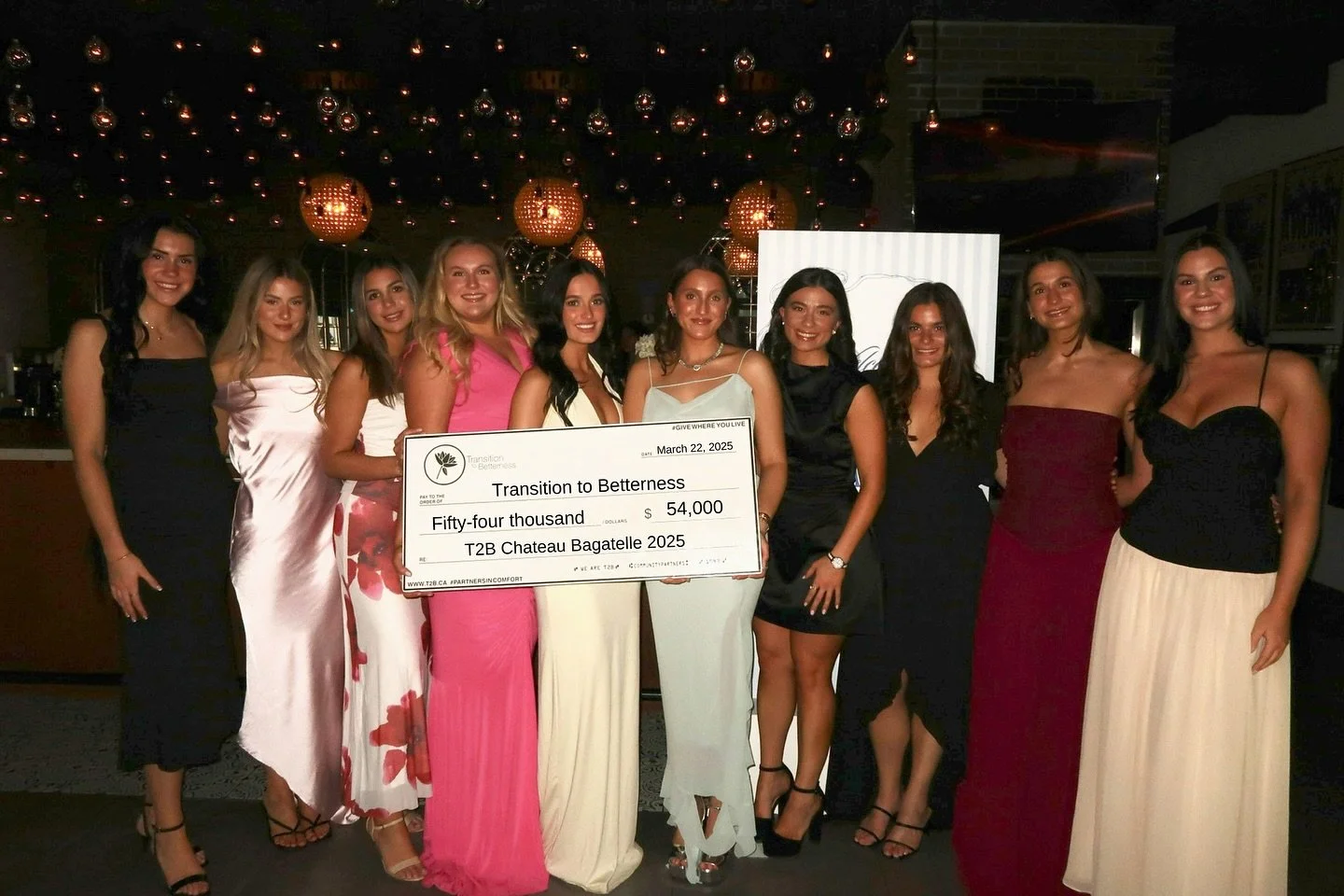 A group of ten young women in formal dresses standing together in a dimly lit venue, holding an oversized check for fifty-four thousand dollars made out to 'Transition to Betterness,' dated March 22, 2025.