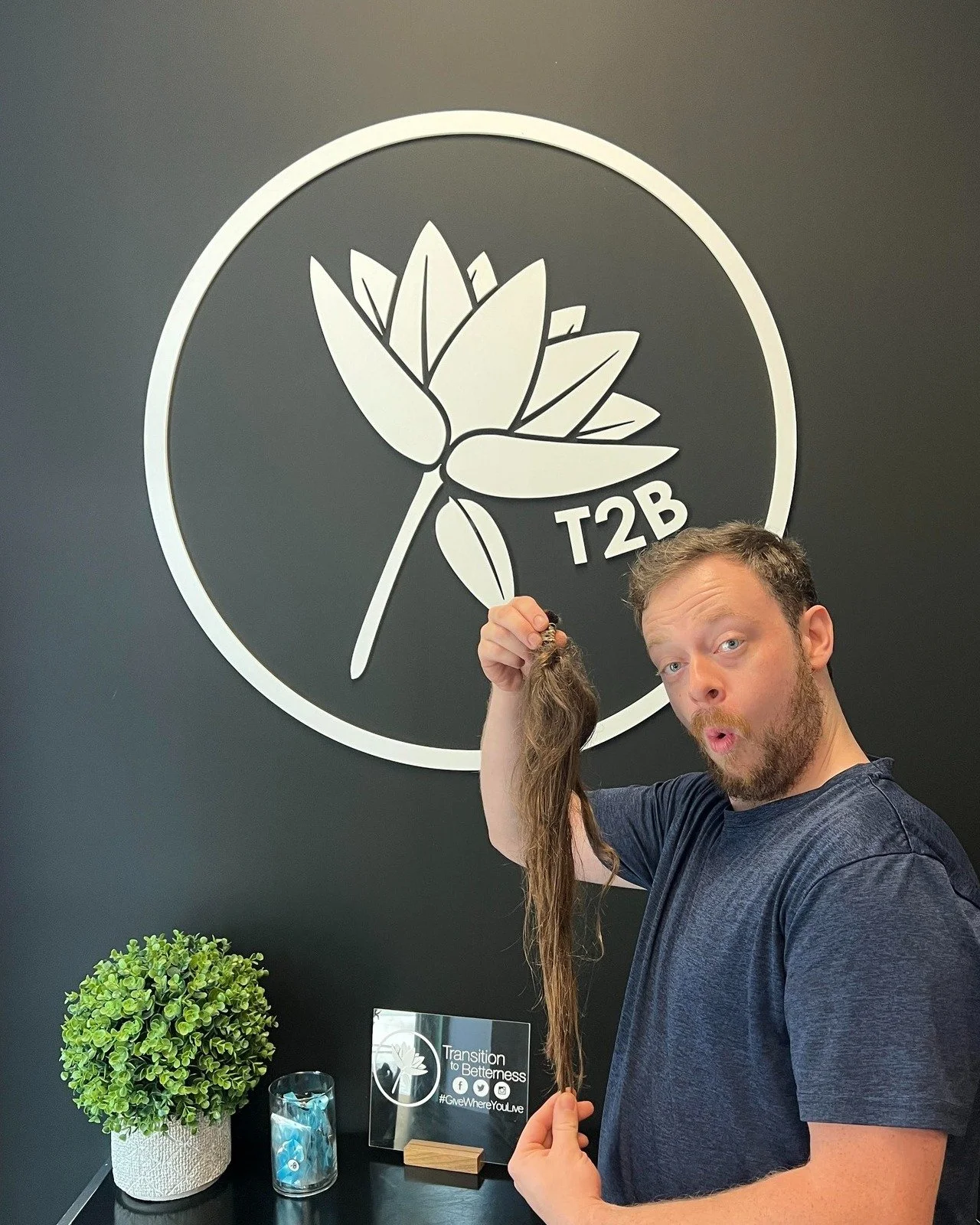 Man holding a long, brown hair piece in front of a wall with a large logo of a lotus flower and 'T2B' text, next to a potted plant and a glass candle holder.