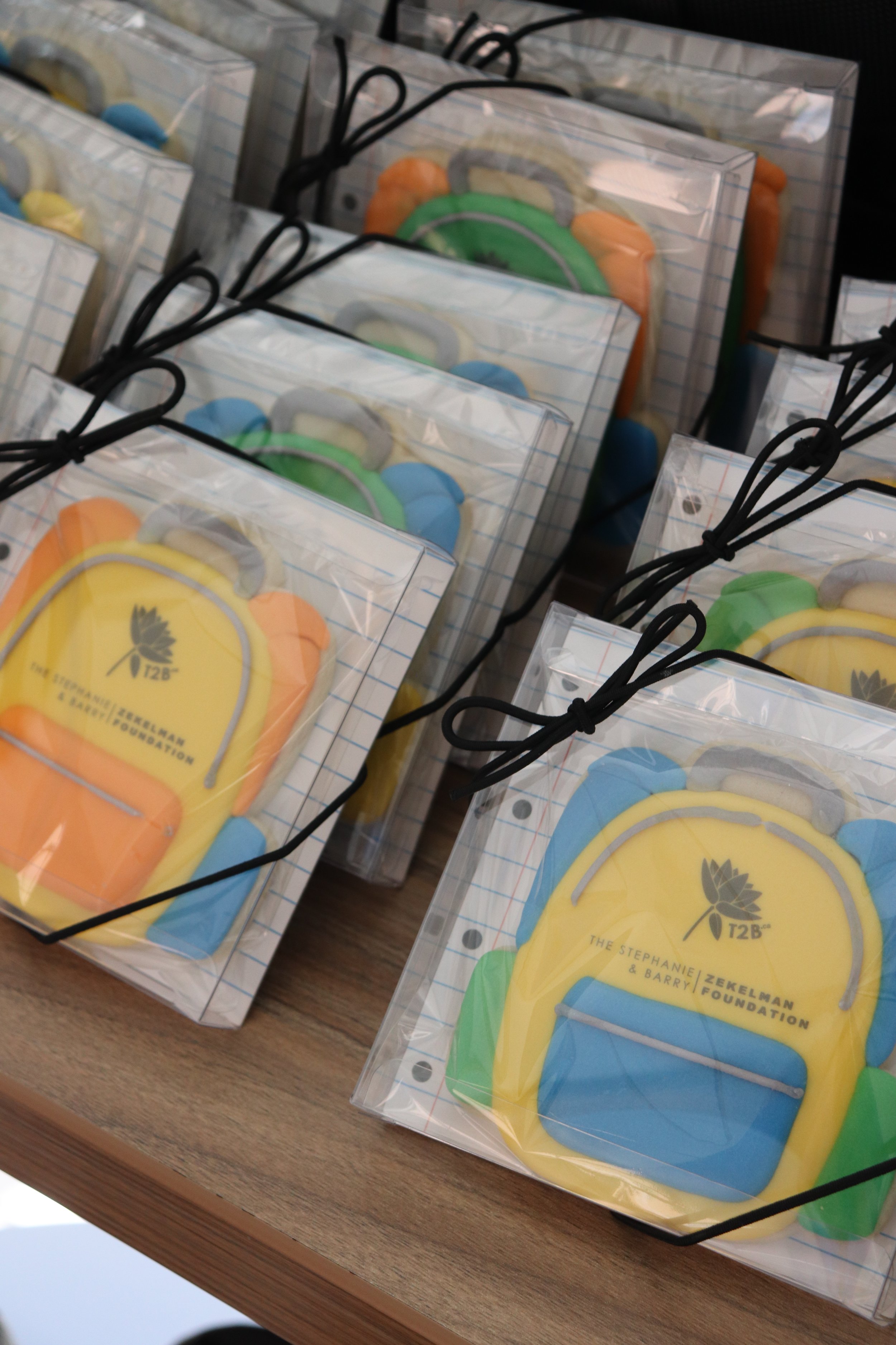 Colorful cookies shaped like backpacks in clear plastic boxes with black bows, on a wooden surface.