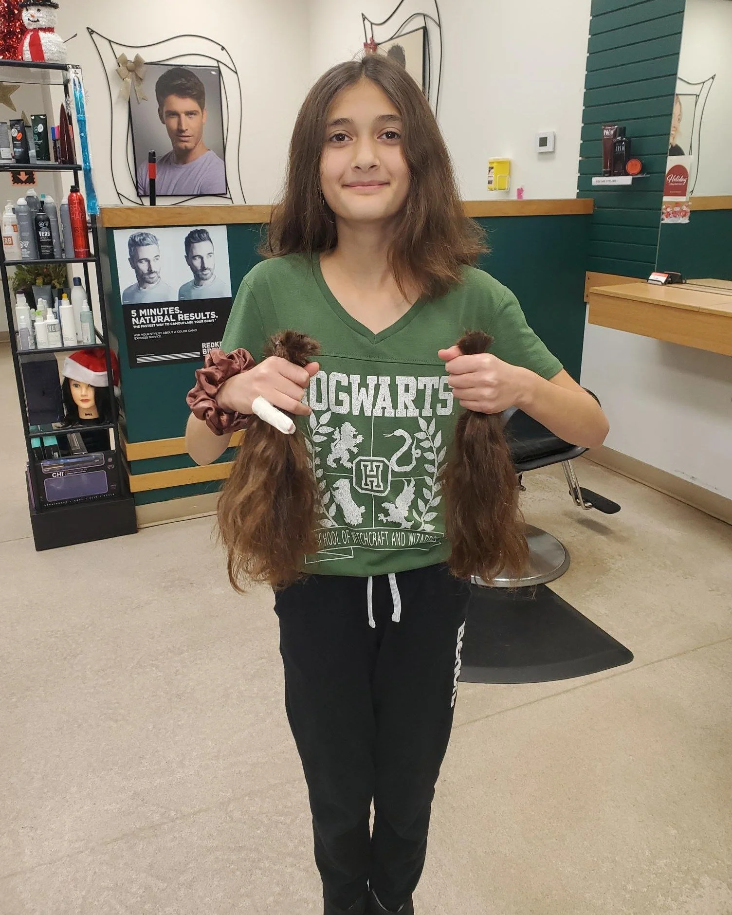 Young girl standing in a hair salon, holding two long curly hair extensions, smiling at the camera. She wears a green Hogwarts t-shirt and black sweatpants.