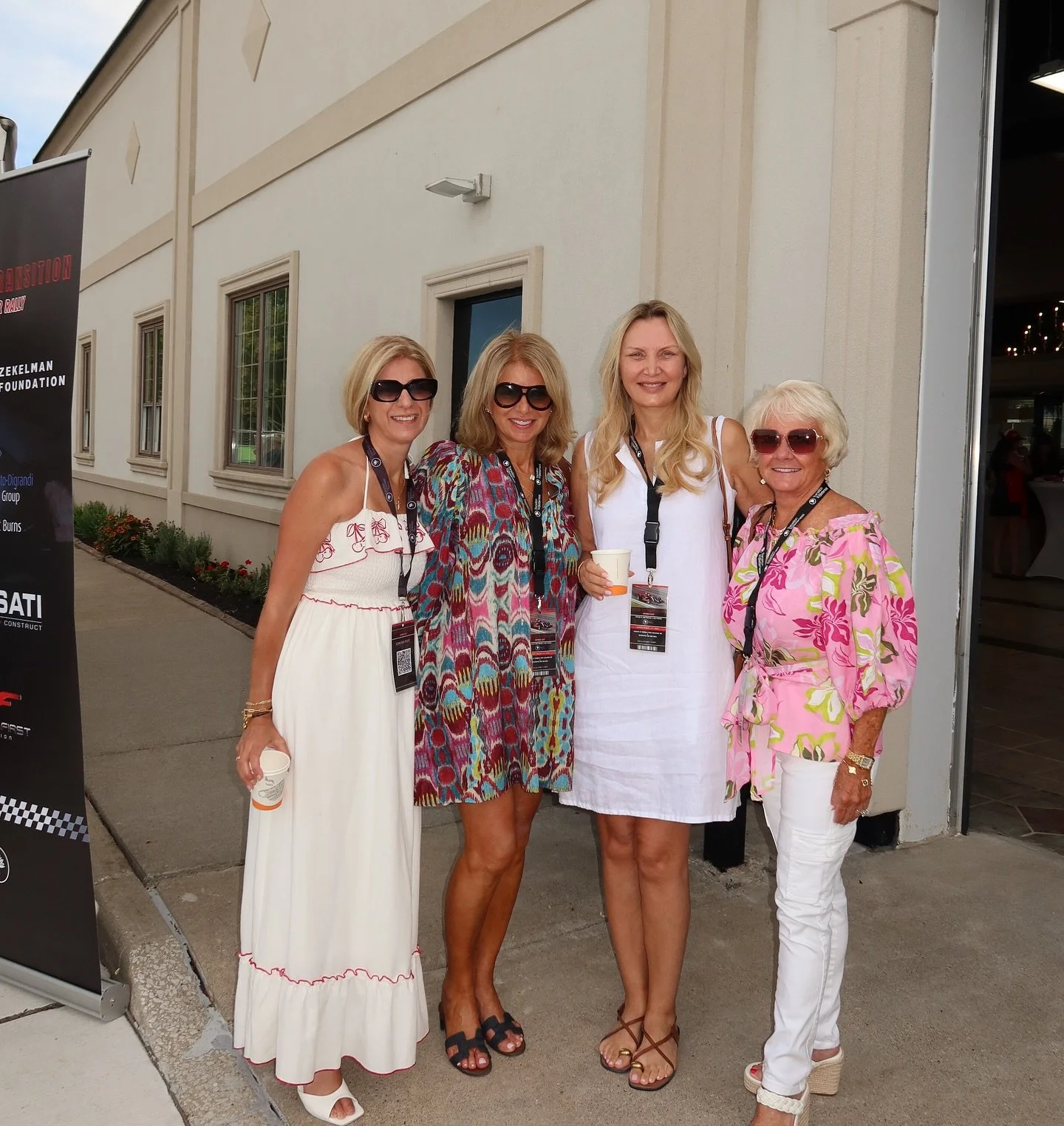 Four women standing together outdoors in front of a light-colored building, smiling. They are wearing summer dresses and sunglasses, holding cups, with name badges around their necks.