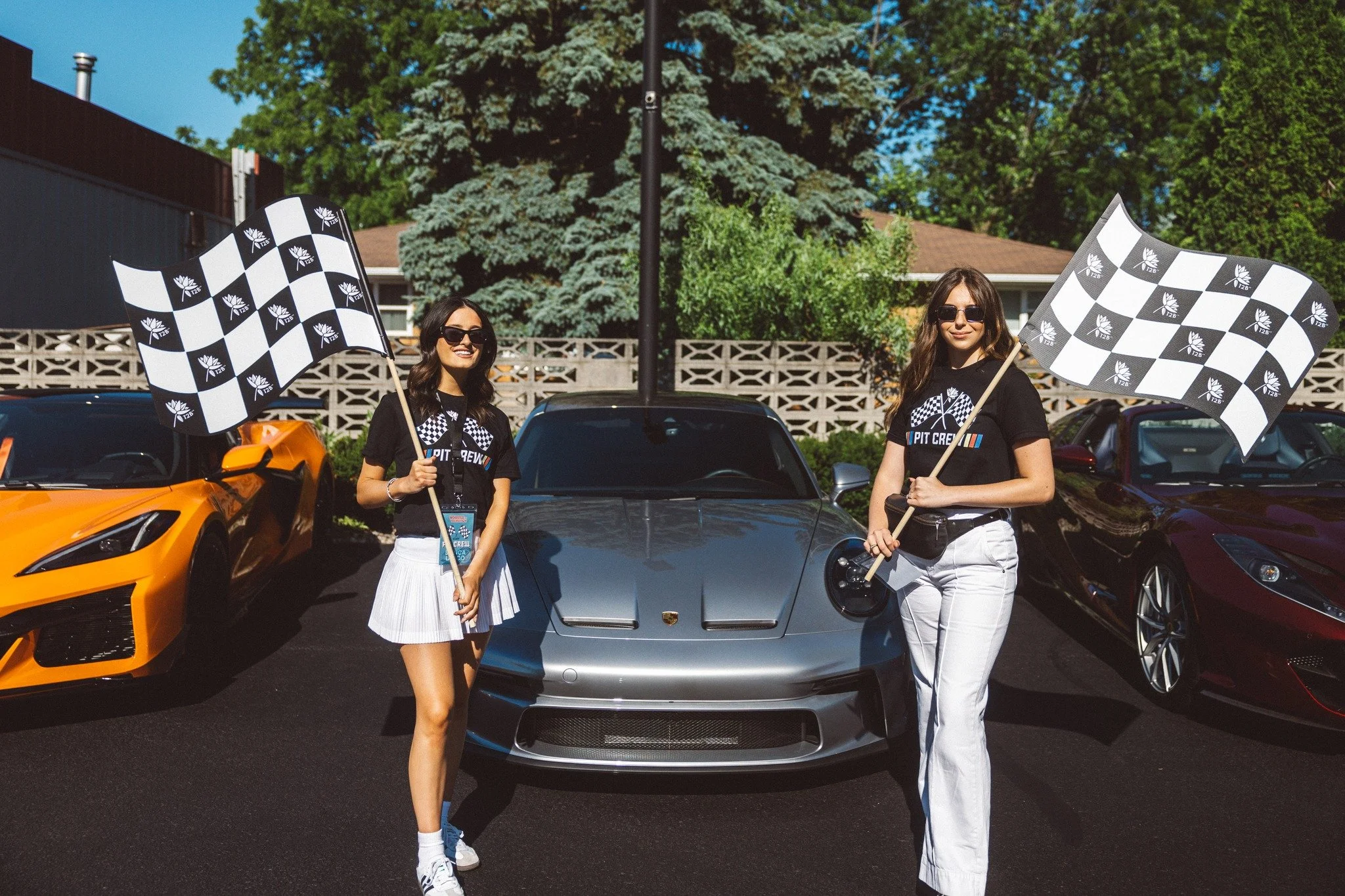 Two women standing in front of sports cars holding checkered flags at a racing event.