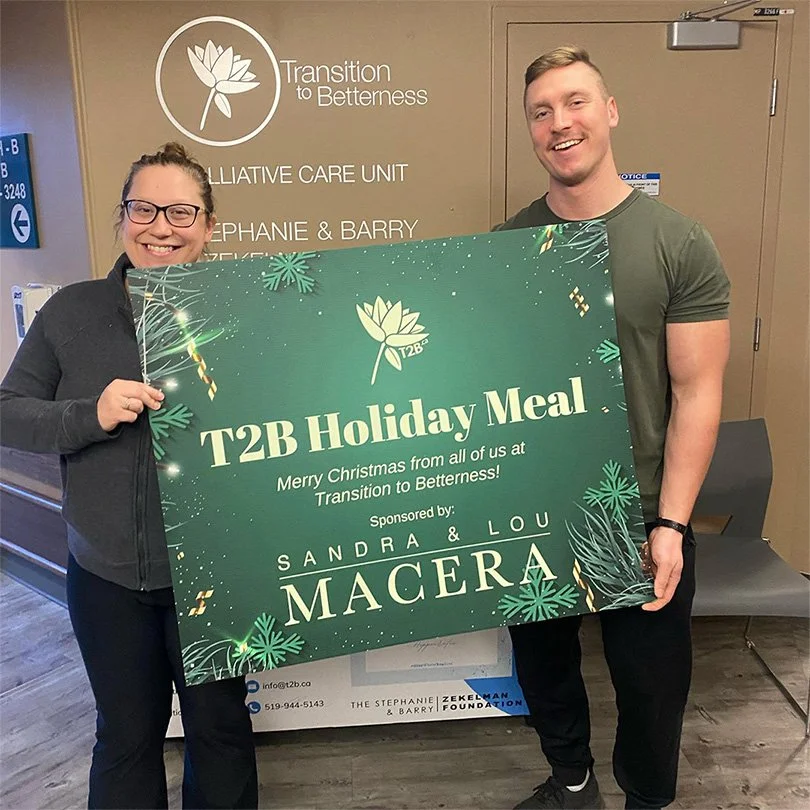Two people smiling and holding a large green holiday meal sign in a healthcare setting. The sign reads "T2B Holiday Meal" and includes Christmas decorations and sponsorship information.