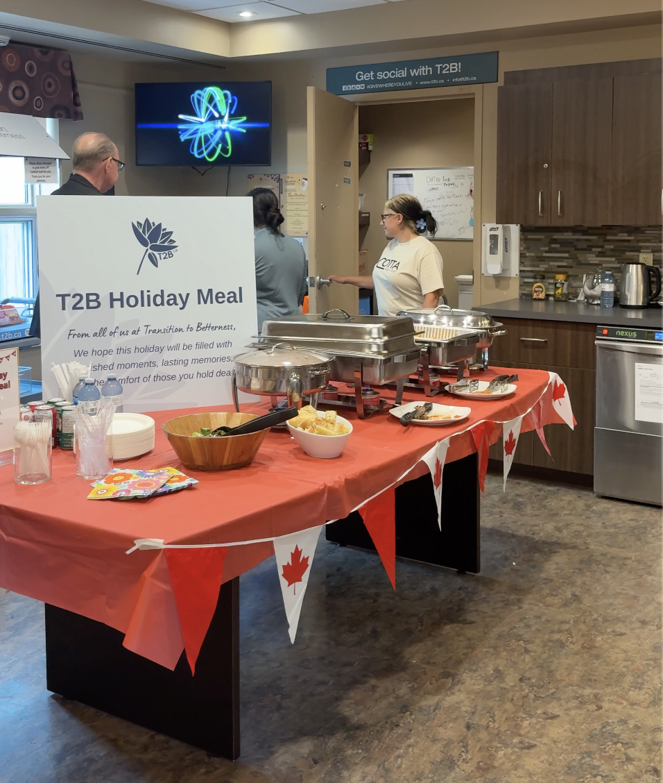 A buffet setup with Canadian flag bunting, serving trays, bowls of food, and people serving themselves, with a T2B Holiday Meal sign on the table, in a room with kitchen appliances and a wall-mounted TV.