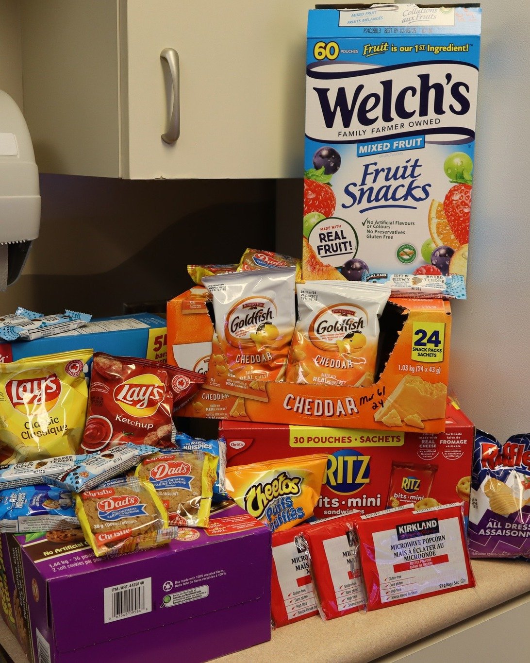 Variety of snack foods, including Welch's mixed fruit snack box, Goldfish cheddar crackers, Lay's classic chips, Dads cookies, Ritz Bits mini sandwiches, Cheetos puffs, Kirkland microwave popcorn, and several small snack packets, arranged on a counter with a cabinet in the background.