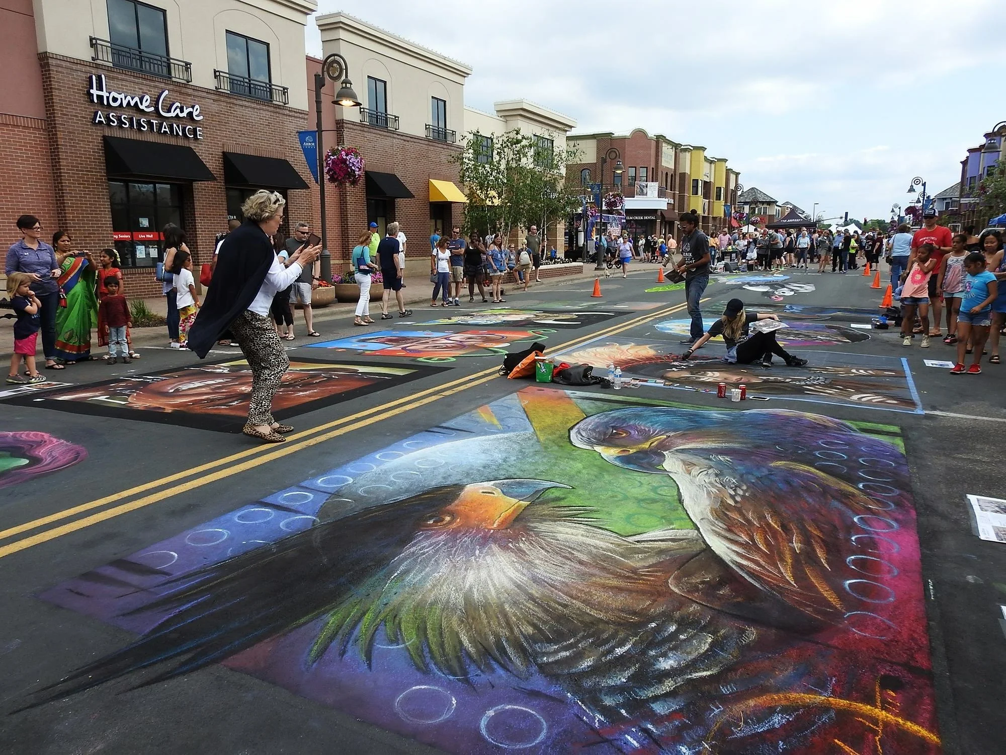 Street art festival with chalk artist creating large, colorful chalk drawings on pavement, including a detailed eagle. Pedestrians and spectators watch, some taking photos, in a bustling town center with shops and buildings in the background.