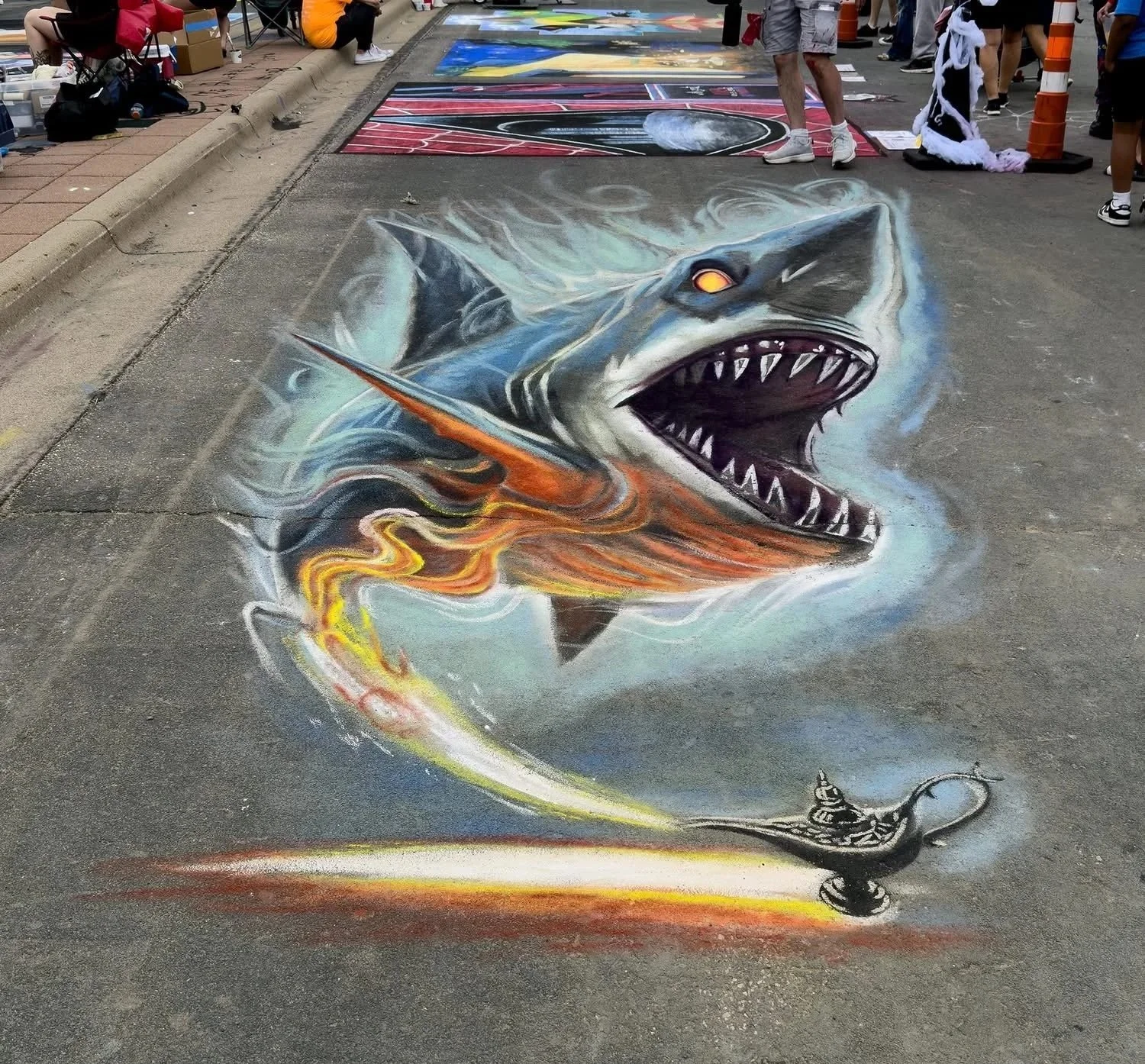 Colorful 3D chalk art of a fierce shark with flames and smoke, chasing a magic lamp, on a street with people and a sidewalk.