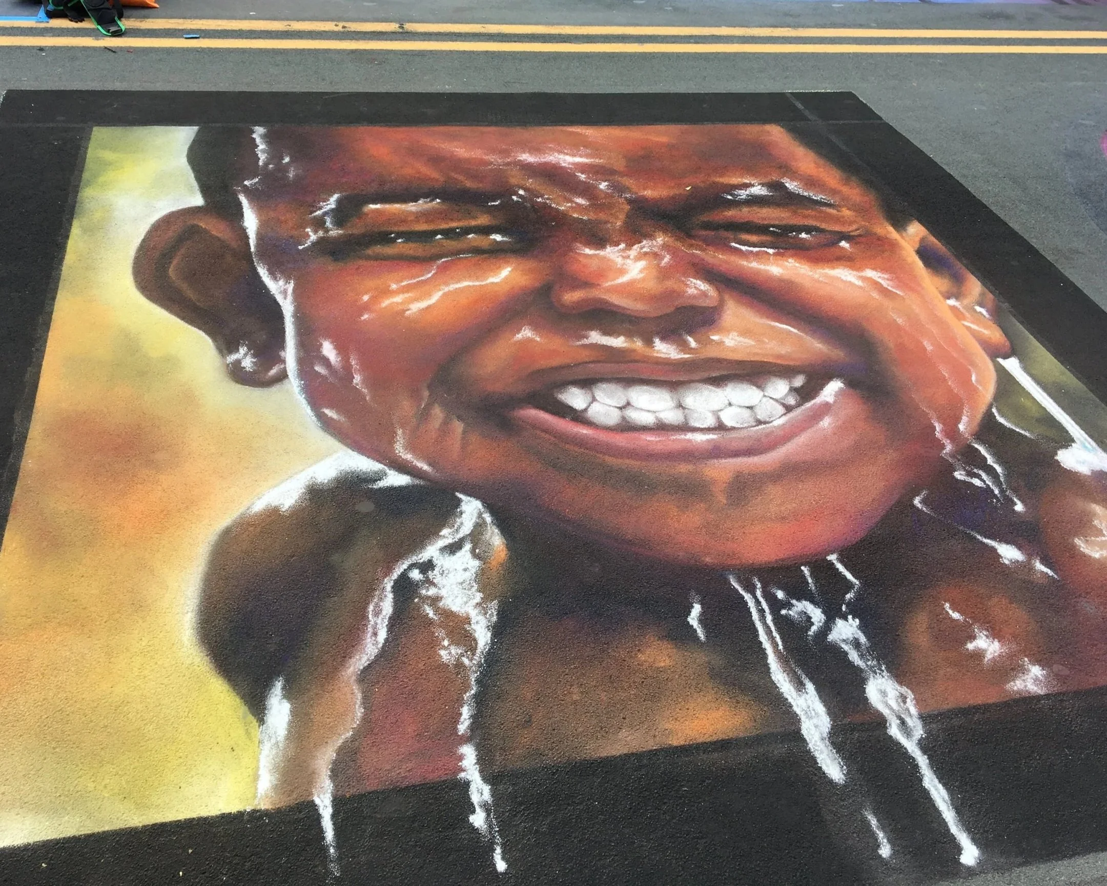 A large, realistic chalk drawing of a smiling child's face on the street, with water or white chalk dripping from the chin and shoulders.