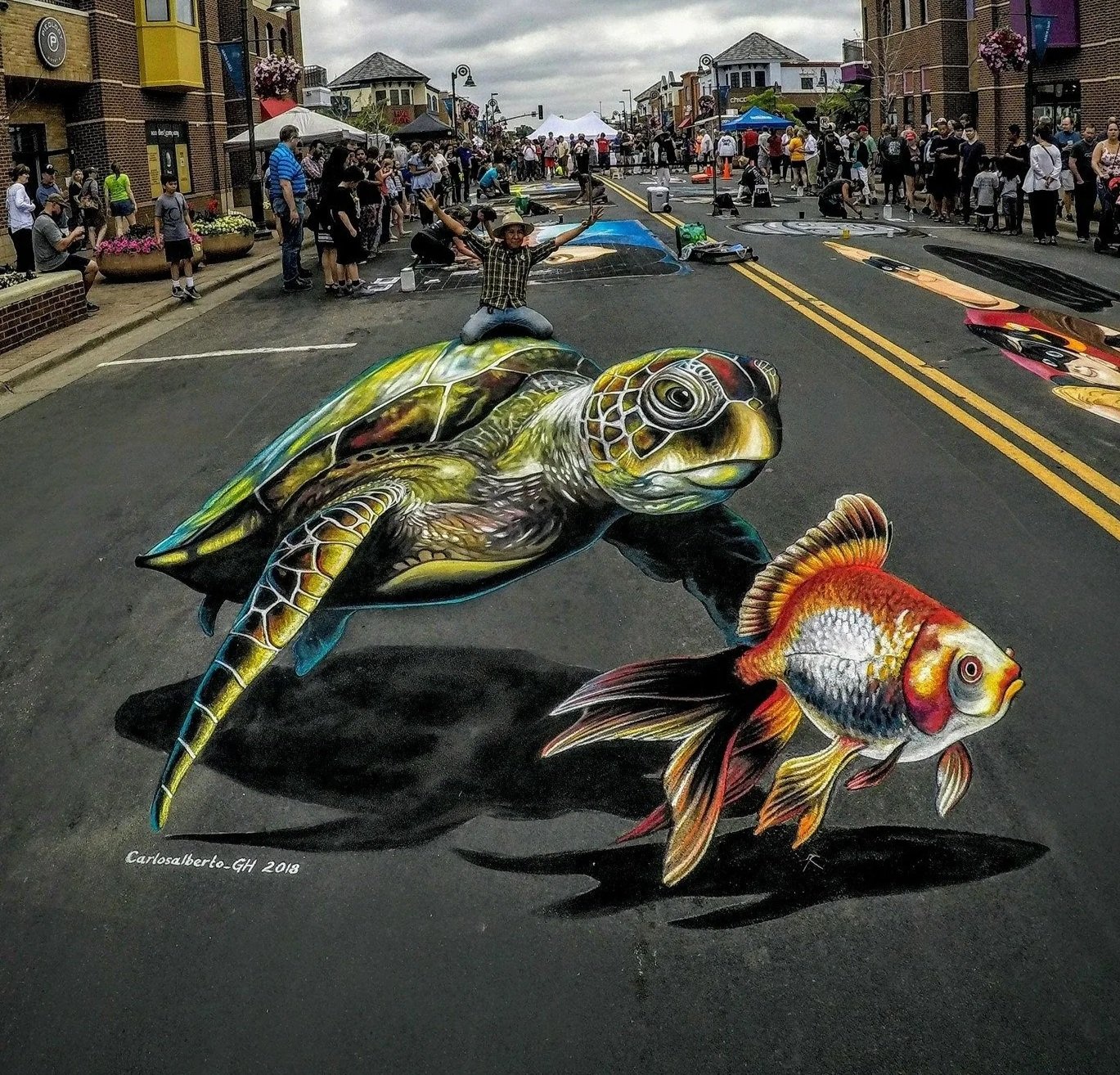 Street scene during a street art festival with a 3D mural of a turtle and fish painted on the street pavement, with people walking and viewing the artwork.