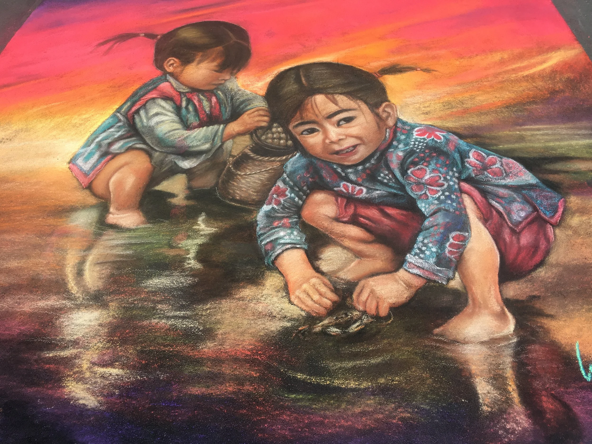 Two young girls in traditional clothing playing in shallow water at sunset, one squatting with a fish, the other leaning forward near a basket.