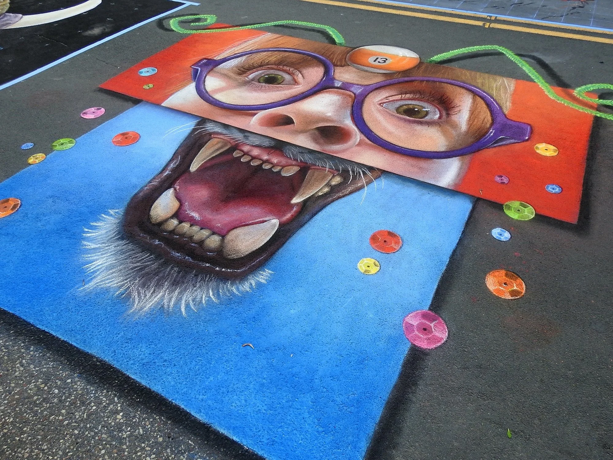 A vibrant chalk art mural on pavement depicting a person with blue eyes, wearing glasses, with a cat face emerging from their open mouth, featuring sharp teeth and pink tongue. Colorful candies are scattered around the artwork.