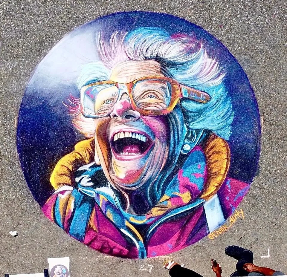 Colorful street art of an elderly woman with gray hair, wearing glasses, laughing with her mouth open, dressed in a jacket with a yellow collar.