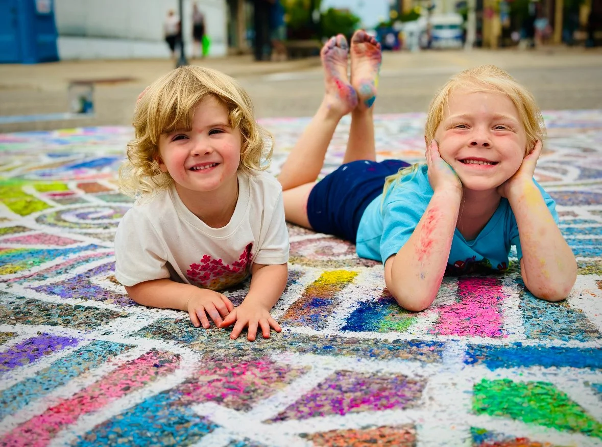 Two smiling children lying on colorful chalk art on pavement in an outdoor urban setting.