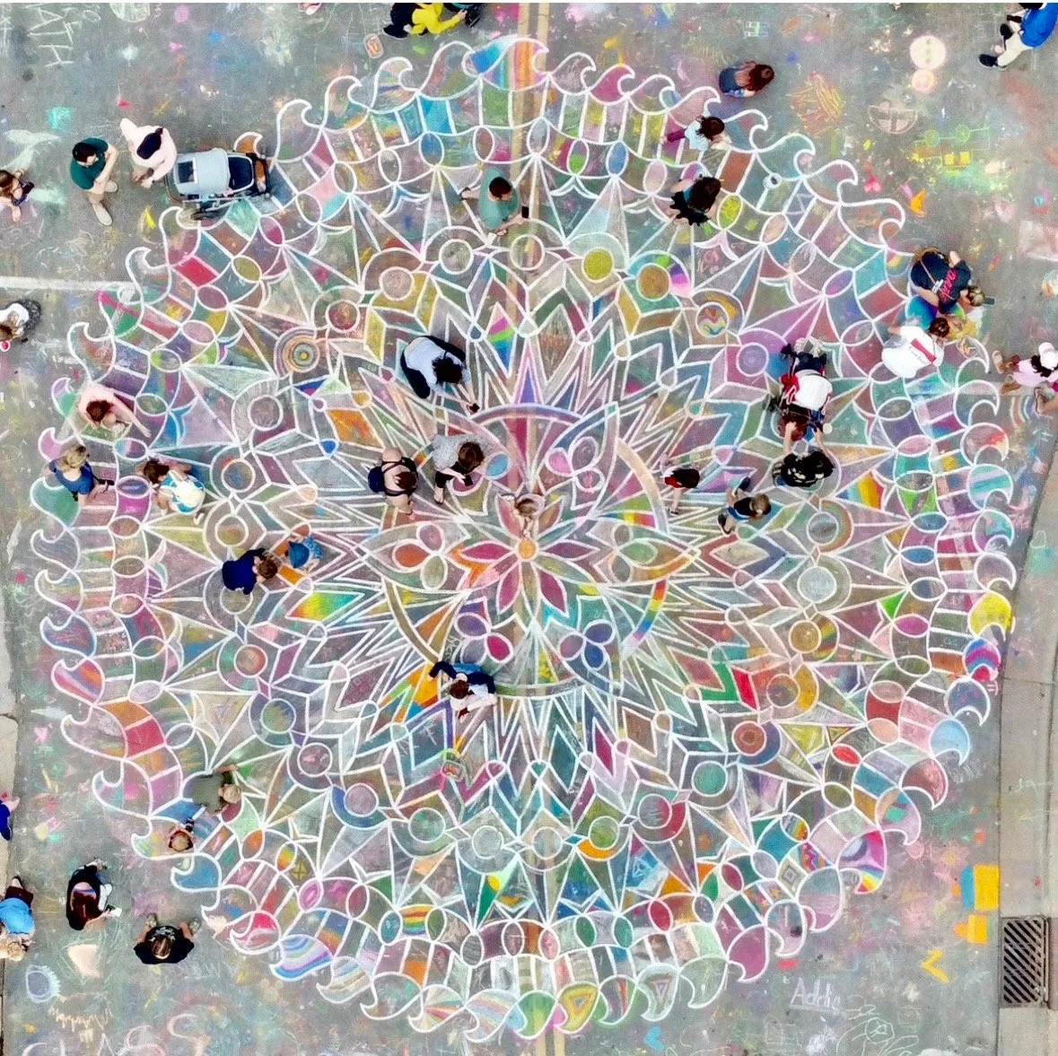 Colorful chalk mandala art on pavement seen from above, with people walking around and through it.