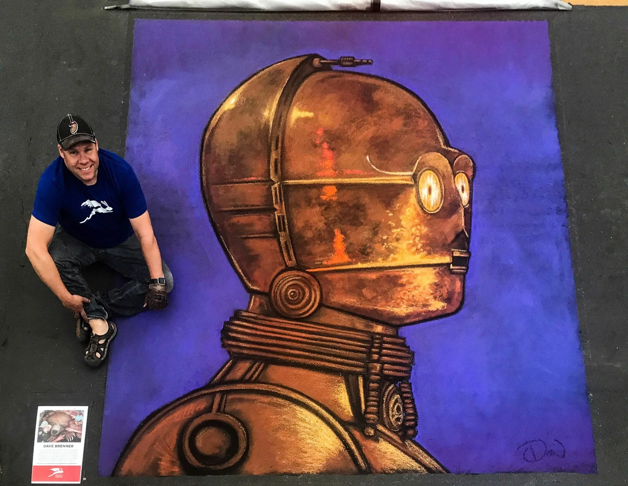 A man crouching next to a large painting of a robot with a brown metallic head and blue background.
