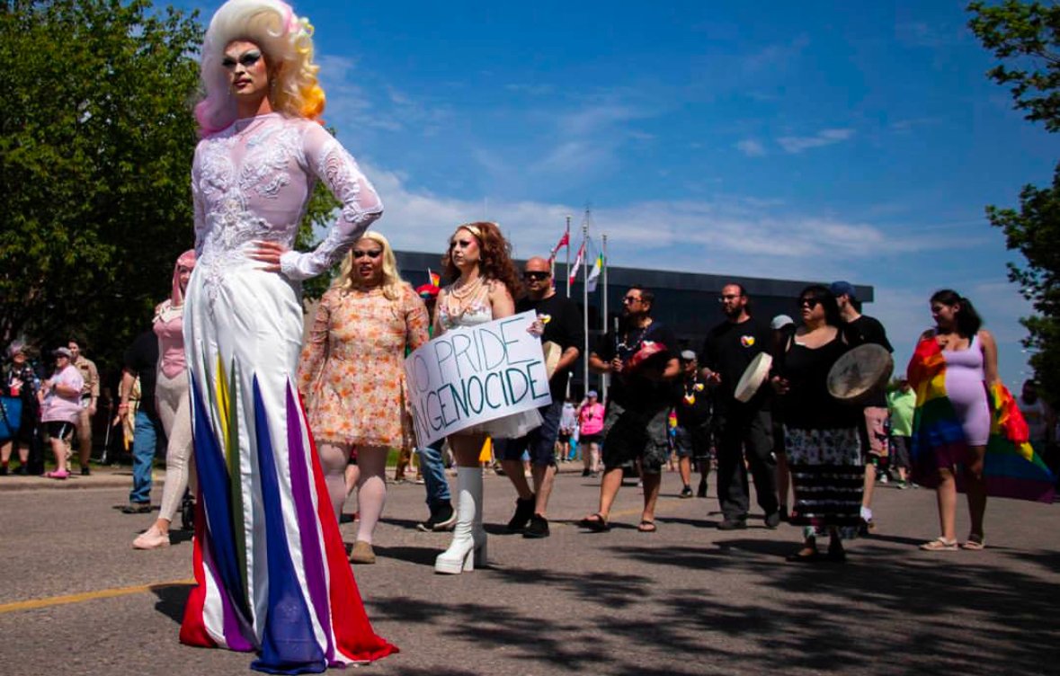 Grand Marshal Flora Hex wears a white long sleeved dress with red, yellow, green, and purple chiffon slits starting below the hip; she wears a platinum blonde wig with the ends dyed with pink, yellow, and orange. March of 2023.
