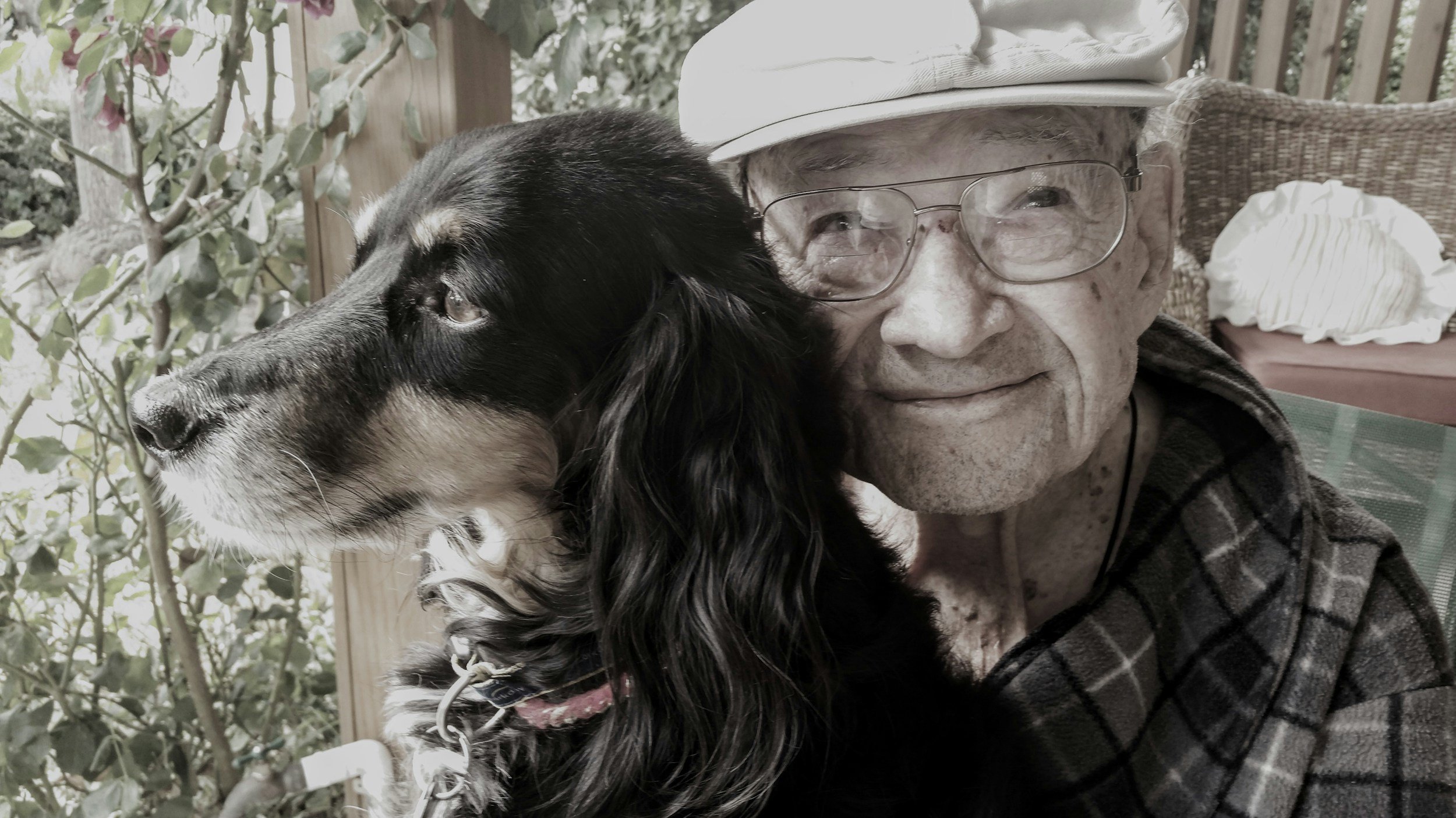 An elderly man with glasses and a white hat smiling as he sits next to a black and white dog with long ears. They are on a porch with greenery and outdoor furniture in the background.