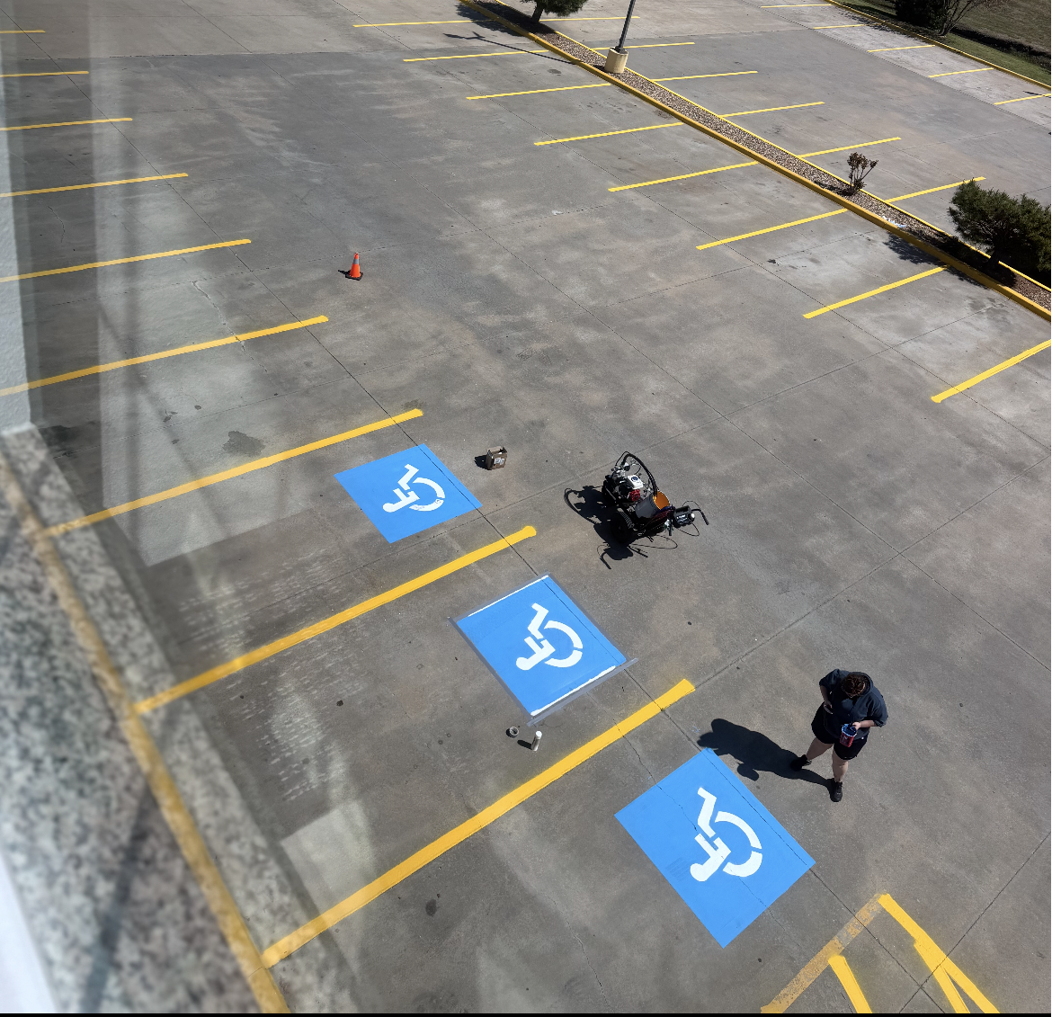 Empty parking lot with a few handicapped spaces being painted, a person holding a paint can, and painting equipment, seen from above.
