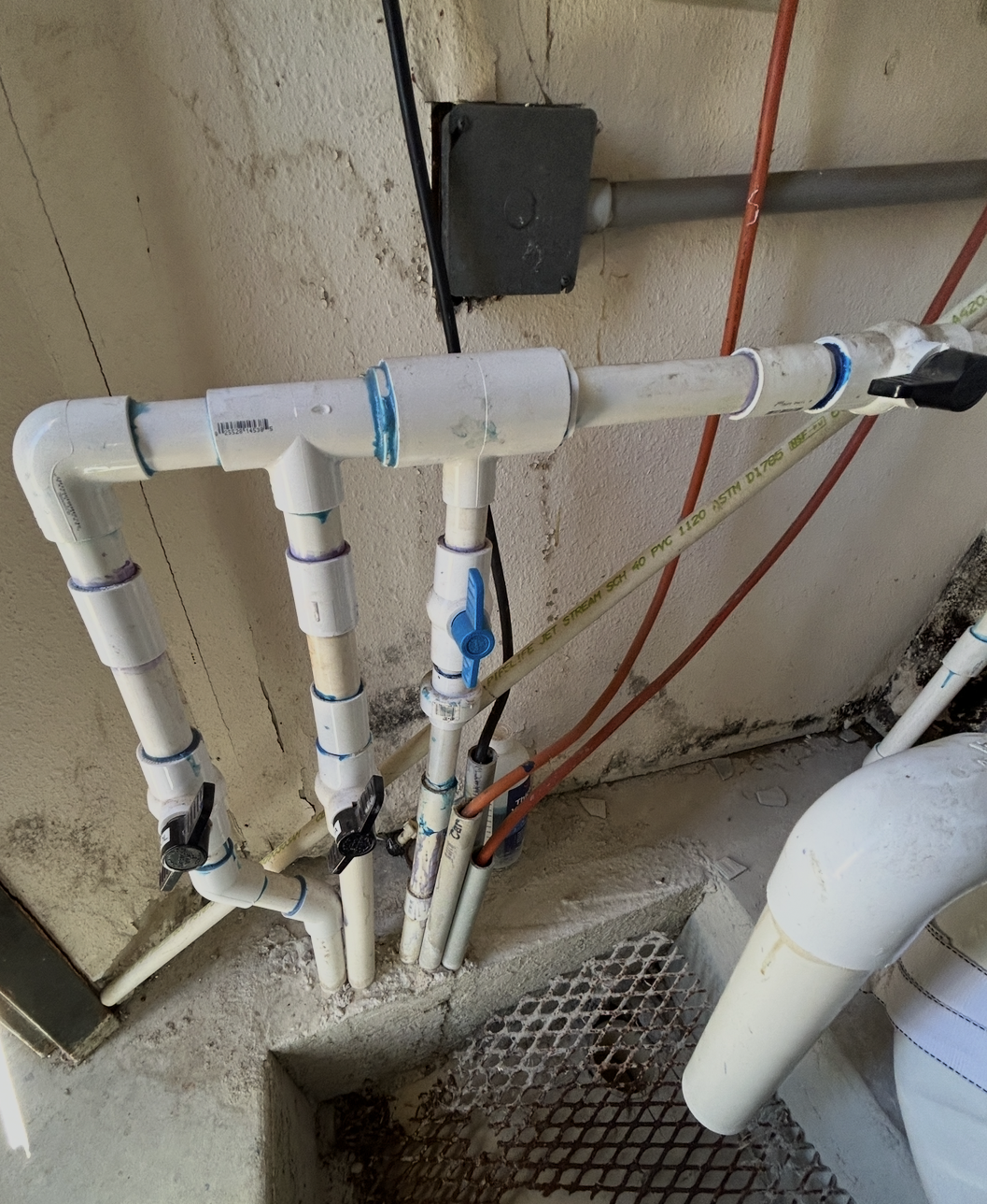 A plumbing system with PVC pipes, valves, and fittings mounted on a wall and floor. Some pipes are connected to a drainage or water system in a utility or mechanical room.
