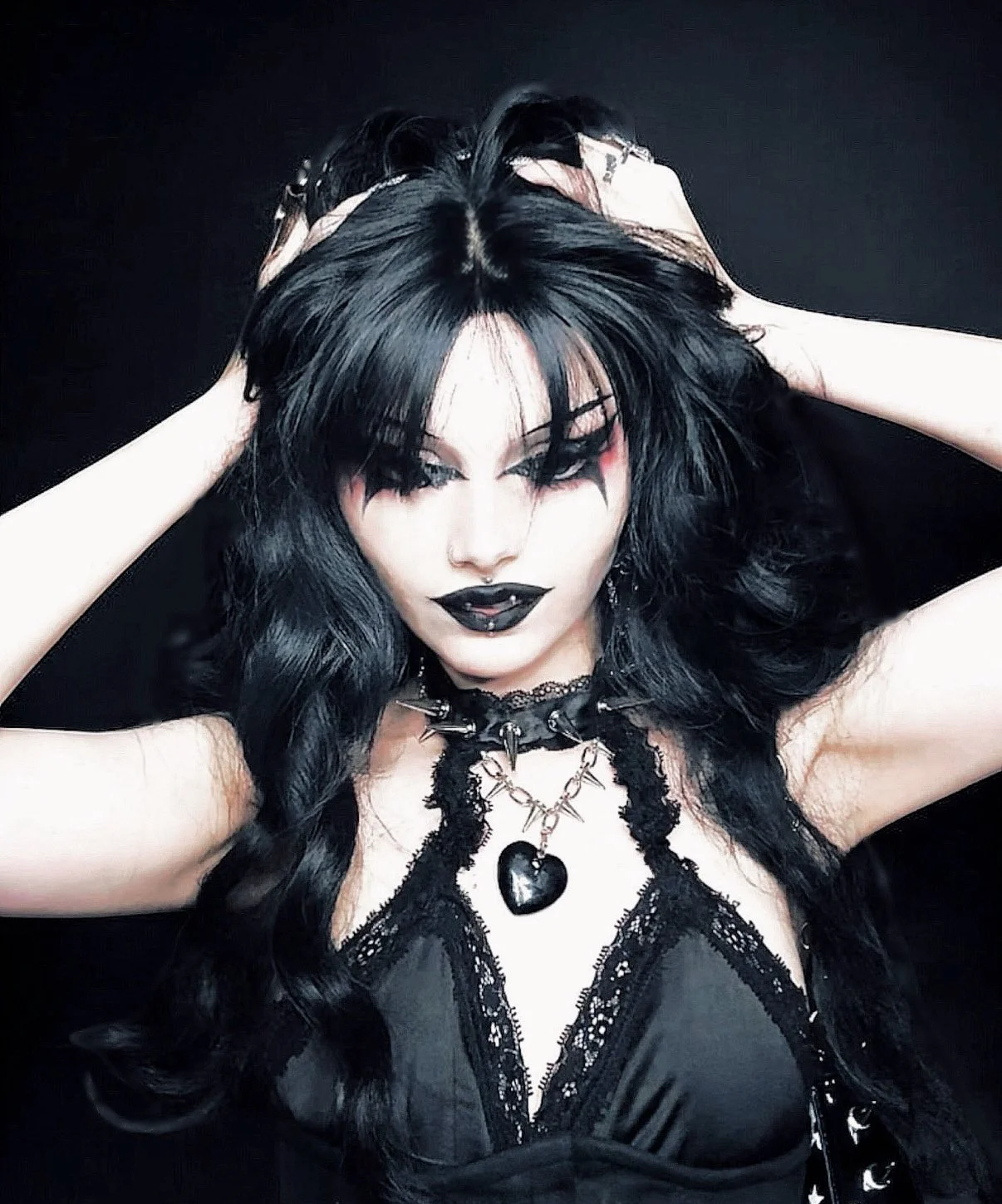 A woman with long black hair, black lipstick, and gothic makeup, holding her hair with both hands, wearing a black lace top, a chokers with spikes and a heart pendant, and dark eye makeup against a dark background.