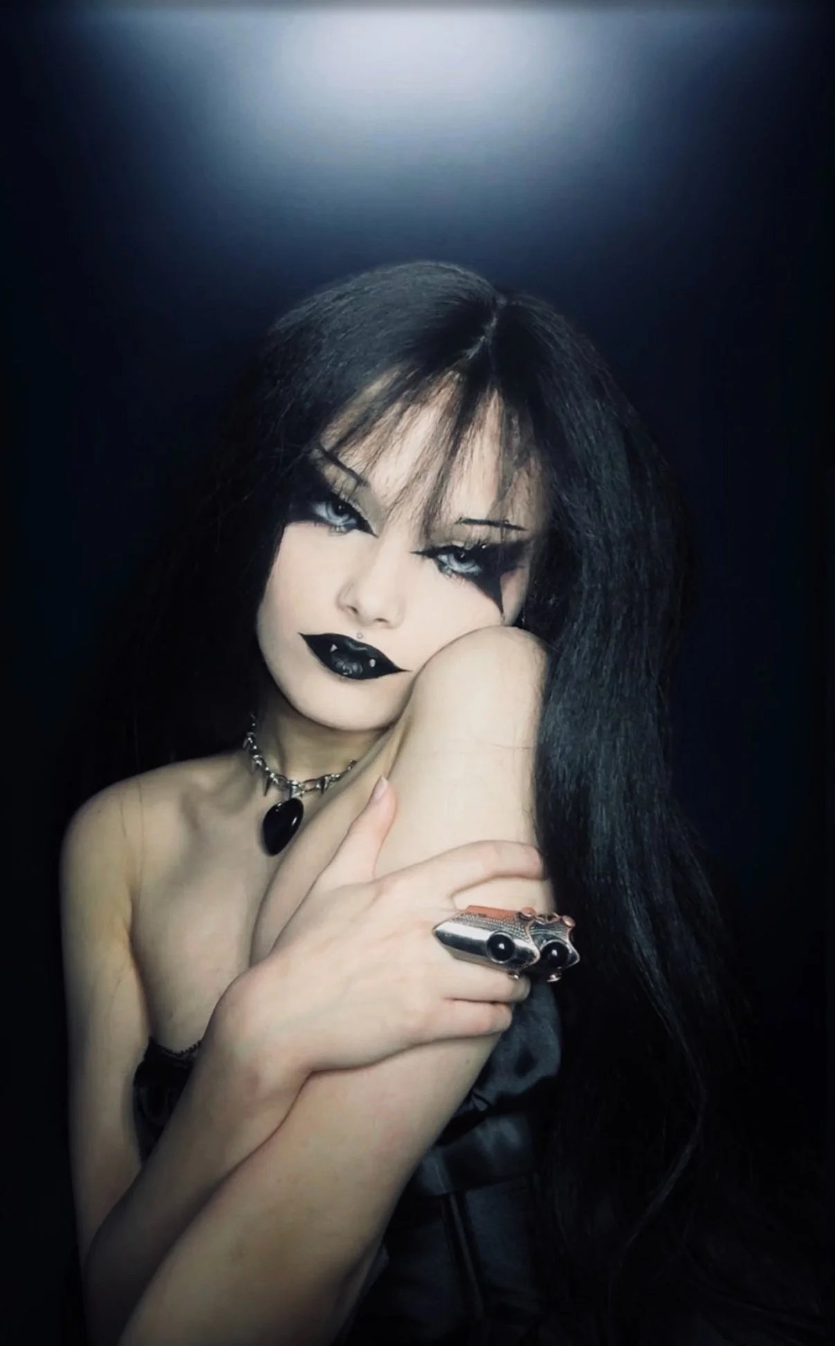 A woman with long black hair and gothic makeup, wearing black lipstick, dark eye makeup, and a choker with a black pendant. She is holding her shoulder and a silver gun-shaped ring.