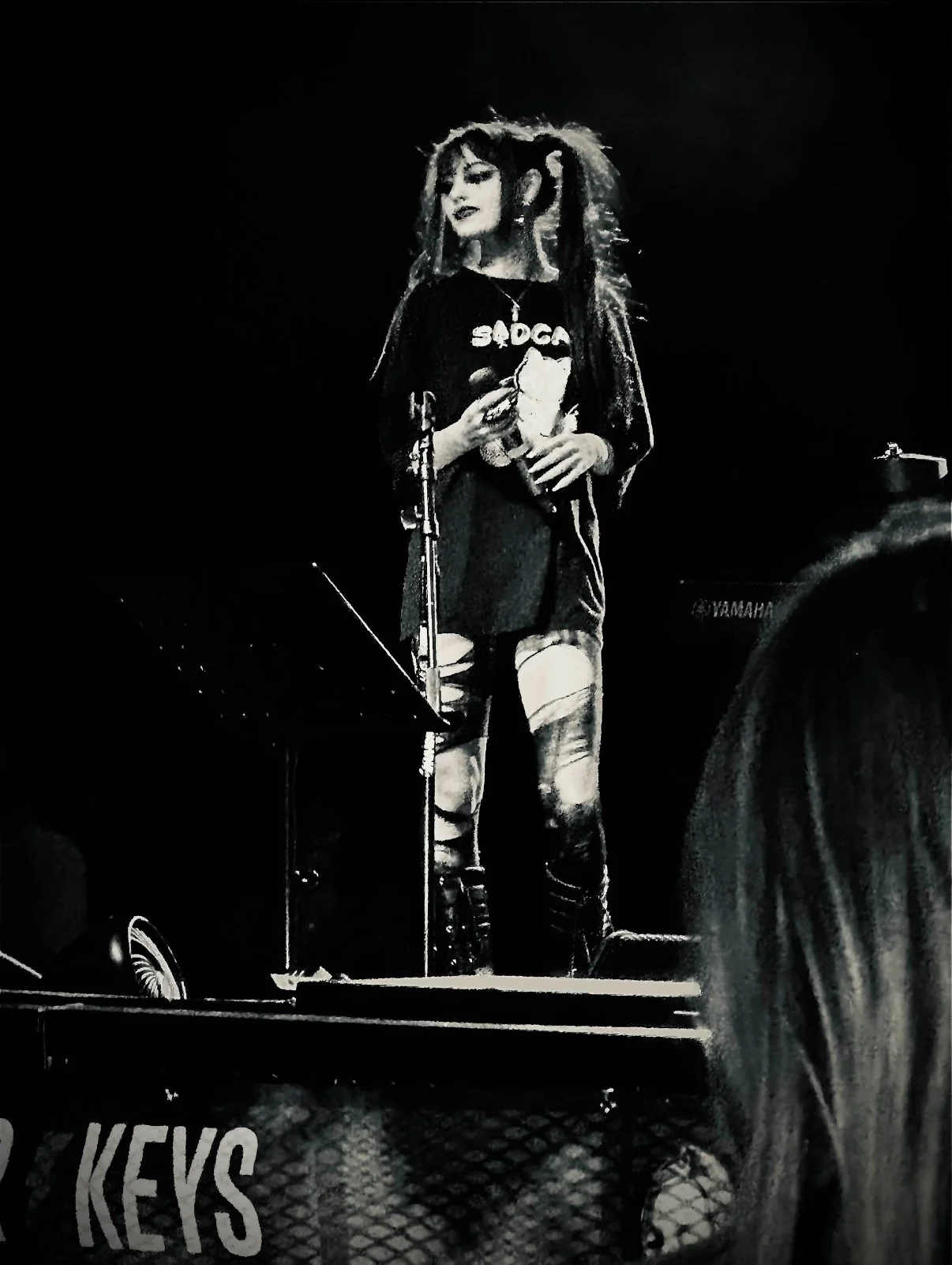 A woman holding a drink standing on stage near a microphone, wearing a black long-sleeve shirt with the word 'BADGIRL' and a cat graphic, ripped tights, and a skirt.