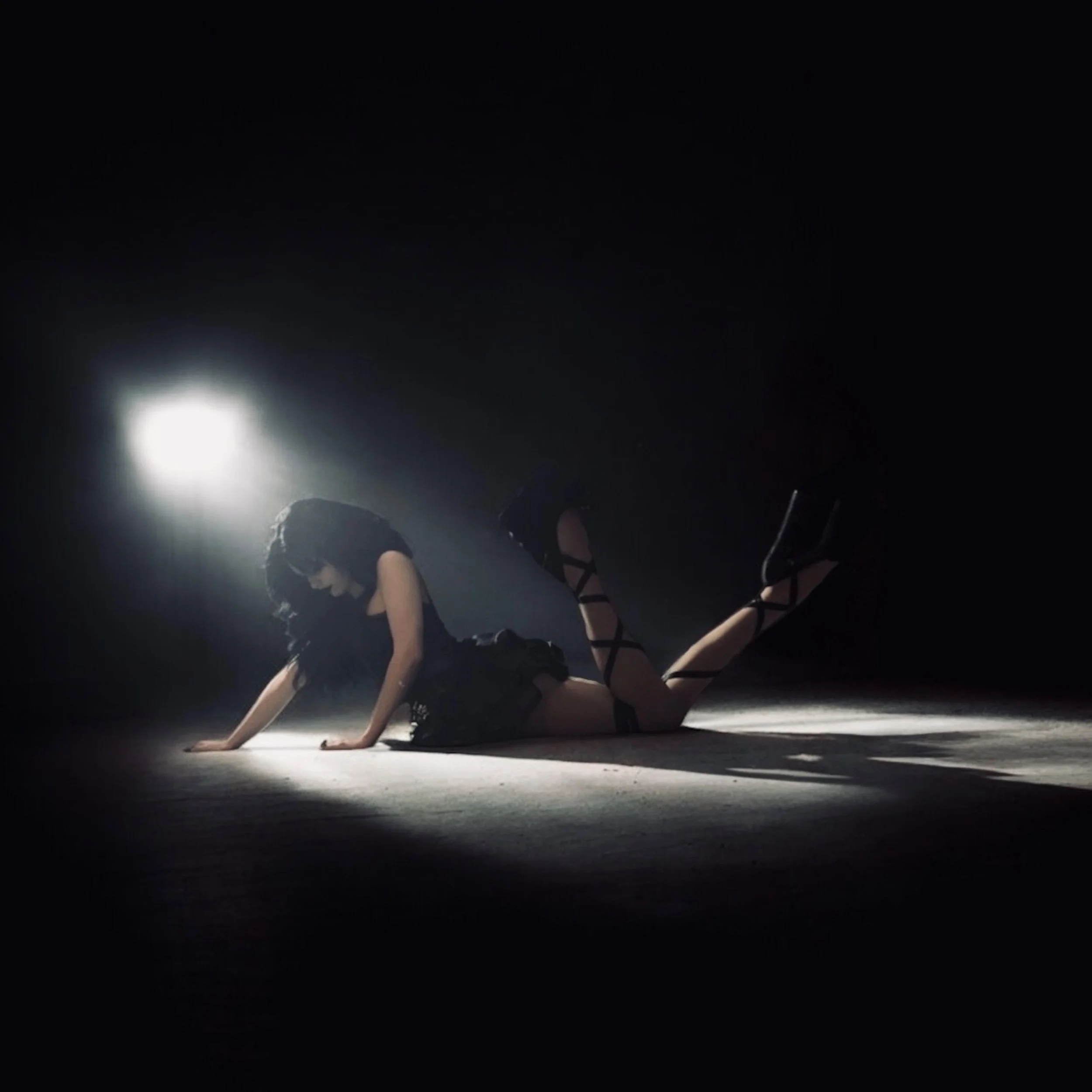 A woman dressed in black with lace-up shoes is posed on the floor, leaning on her hands with her legs bent upwards, illuminated by a spotlight against a dark background.