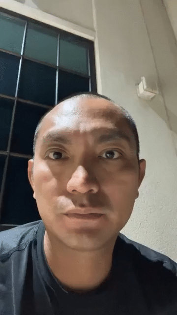 A man taking a selfie indoors, with a window and a beige wall in the background.