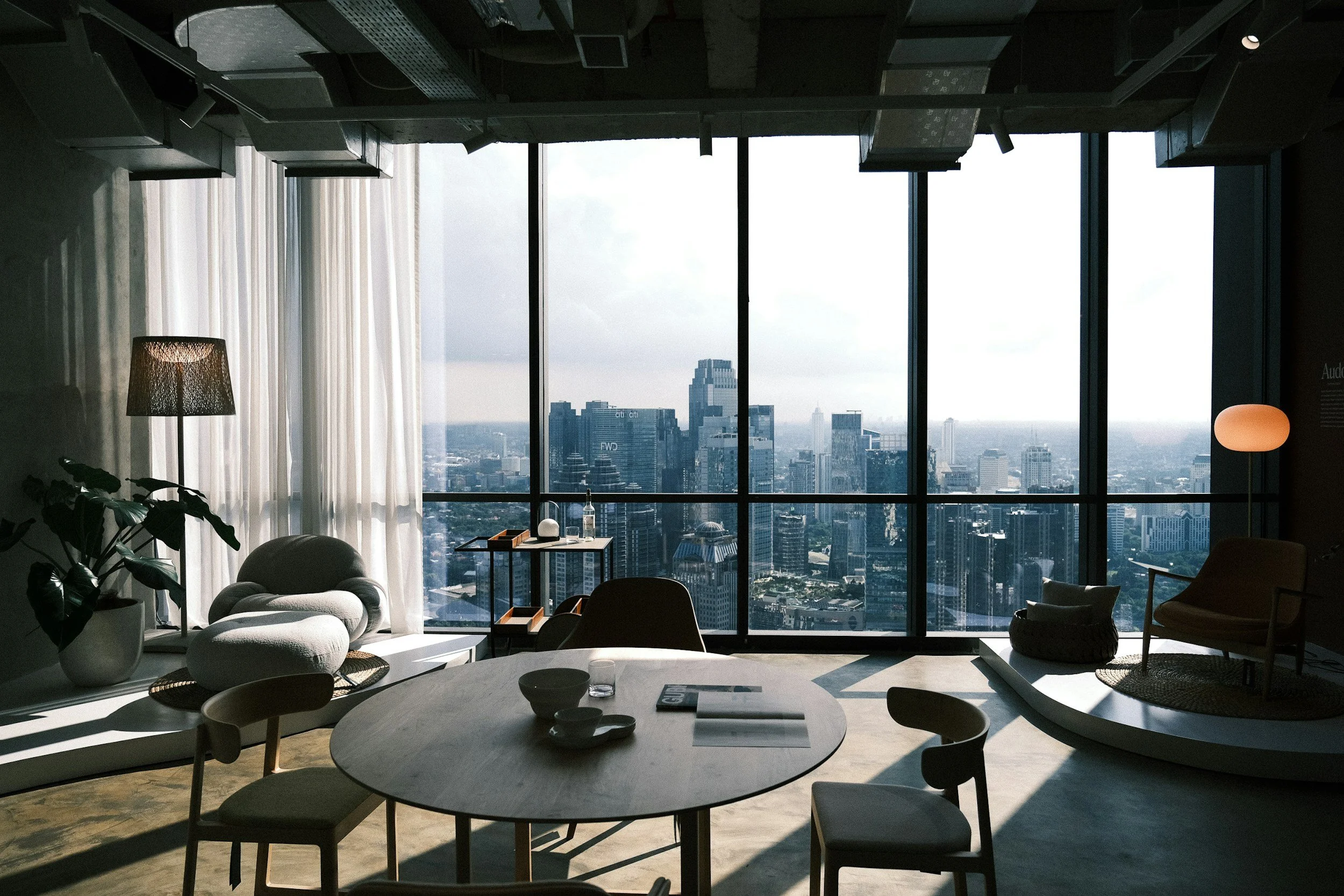 Interior of a modern high-rise apartment with a large floor-to-ceiling window showing a city skyline. The room contains a round dining table with chairs, a cozy lounge area with a chair and ottoman, and decorative lighting.