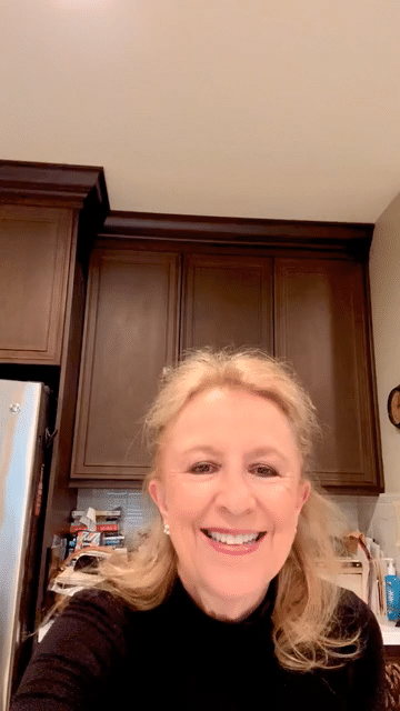 A woman with blonde hair smiling in a kitchen with dark wooden cabinets and a stainless steel refrigerator.