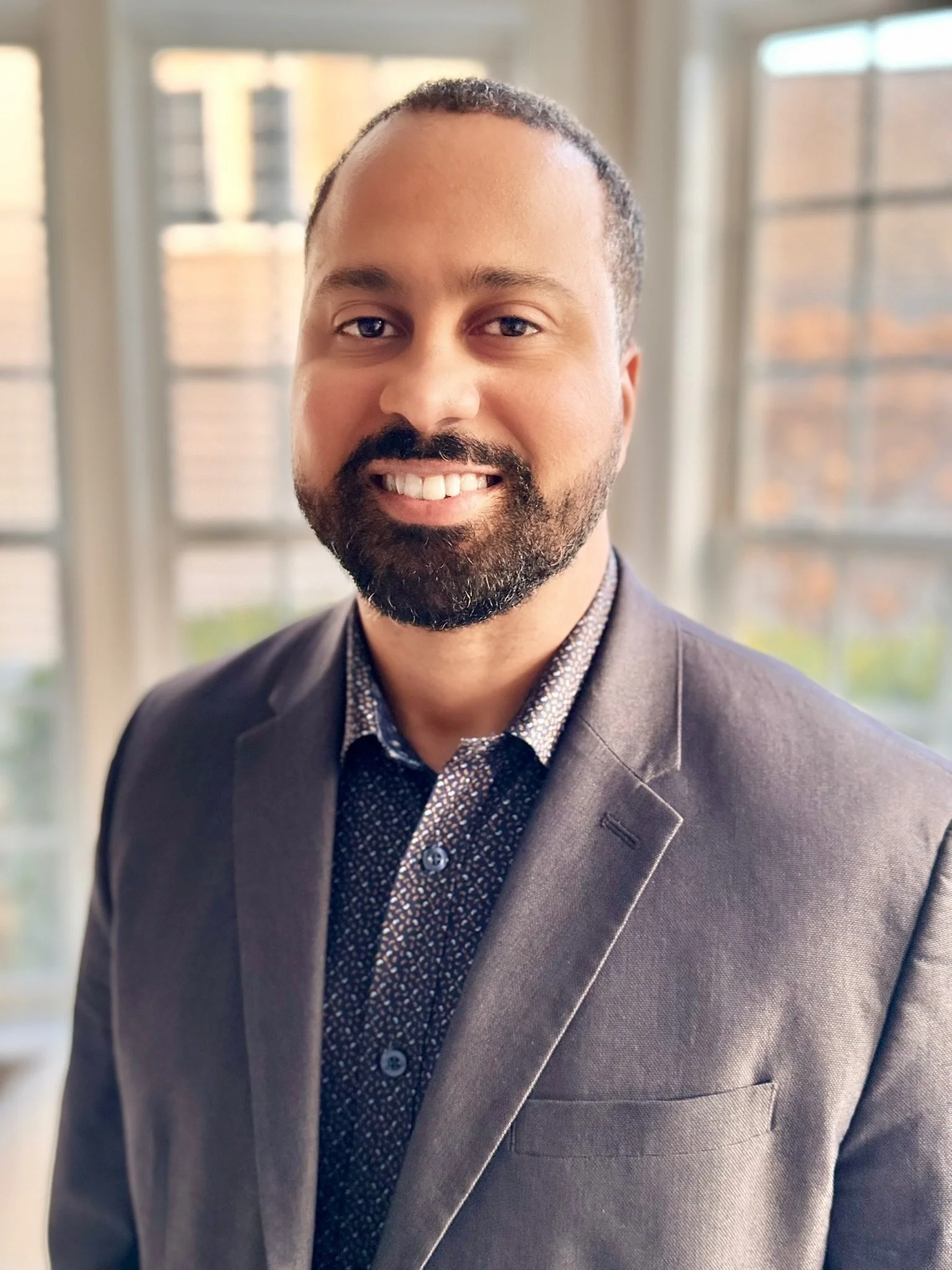 A smiling man with a dark beard and short hair, wearing a dark suit jacket over a patterned shirt, standing in front of large windows with a cityscape in the background.