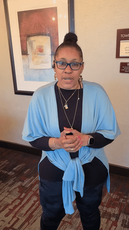 Woman wearing glasses, black shirt, and light blue jacket tied around waist, standing in an indoor setting with artwork and signs on the wall.