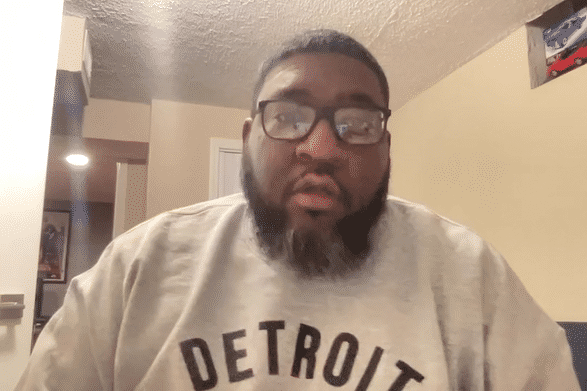 A man with glasses and a beard, wearing a gray t-shirt, sitting indoors.