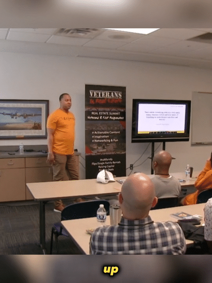 A man in an orange shirt standing in front of a room giving a presentation to seated attendees. There is a screen displaying a slide and a VETERANS banner behind him.