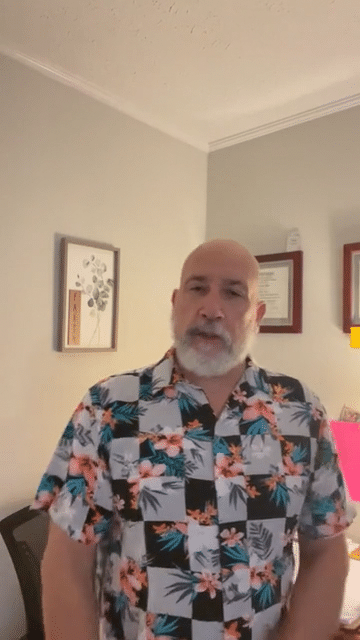 A man with a beard and mustache wearing a colorful Hawaiian shirt standing indoors with framed artwork and certificates on the wall behind him.