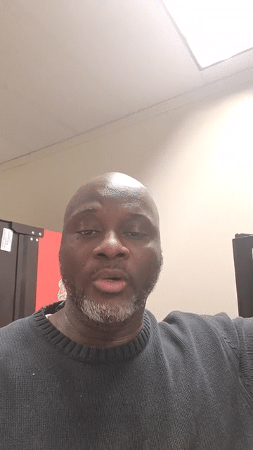 A man taking a selfie indoors, with a beige wall, ceiling, and black lockers in the background.