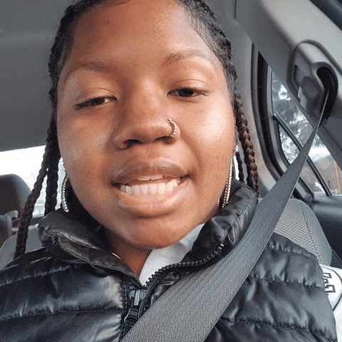 Close-up of a young woman with braids wearing a black jacket and seatbelt inside a vehicle.